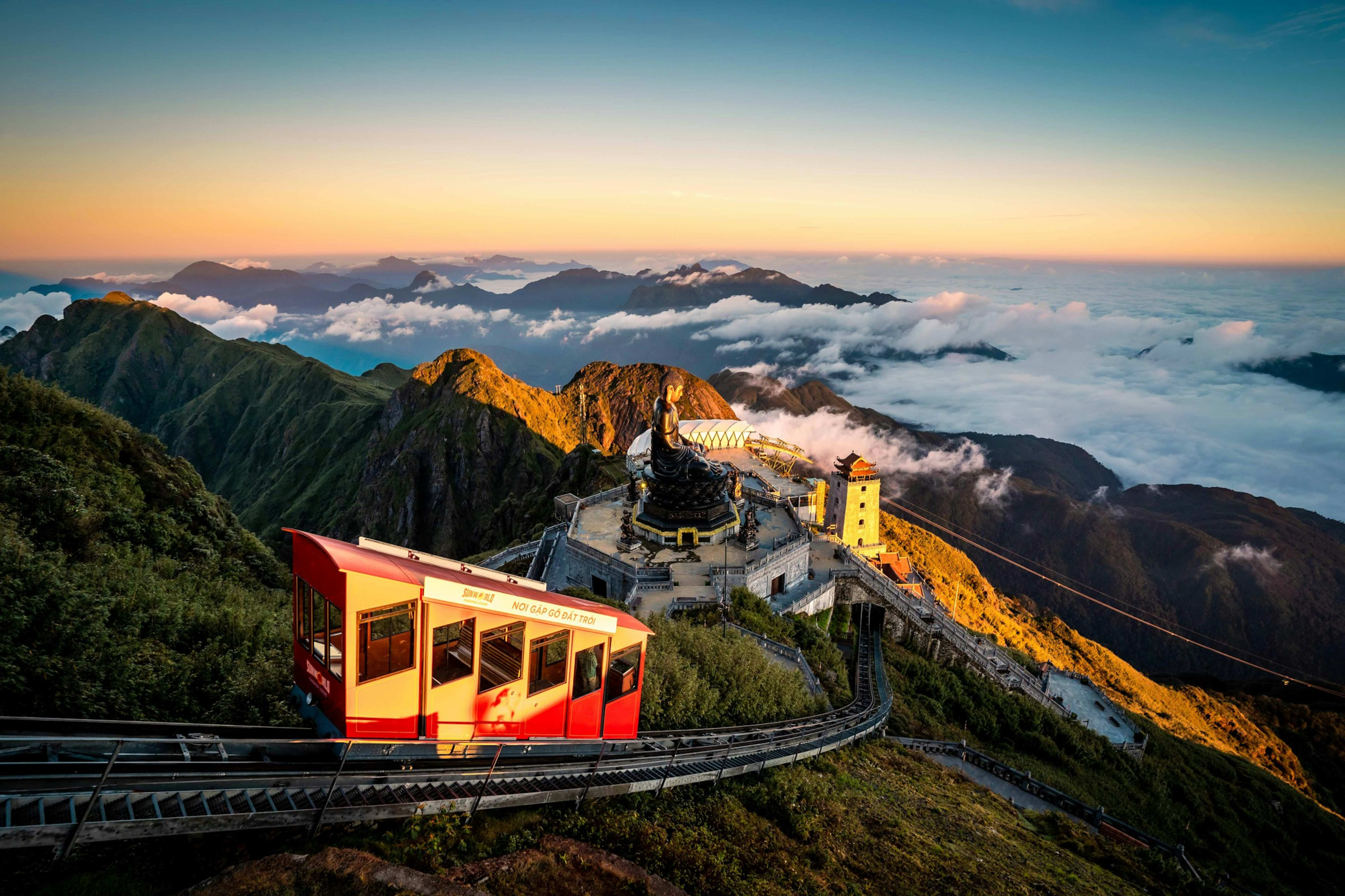 The red funicular carriage climbing the last ridge to the Fansipan summit at sunrise, a bronze Buddha and clock tower standing above a sea of cloud.
