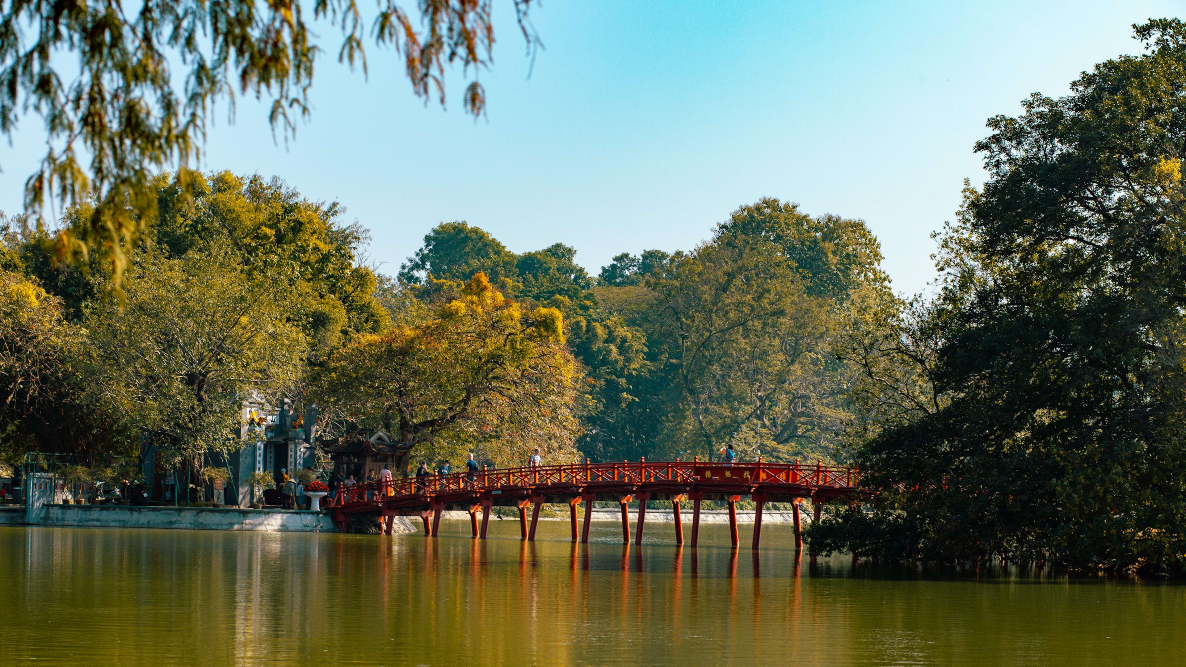The red wooden Húc Bridge seen across Hoàn Kiếm Lake, framed by leaning trees and doubled in the still green water.