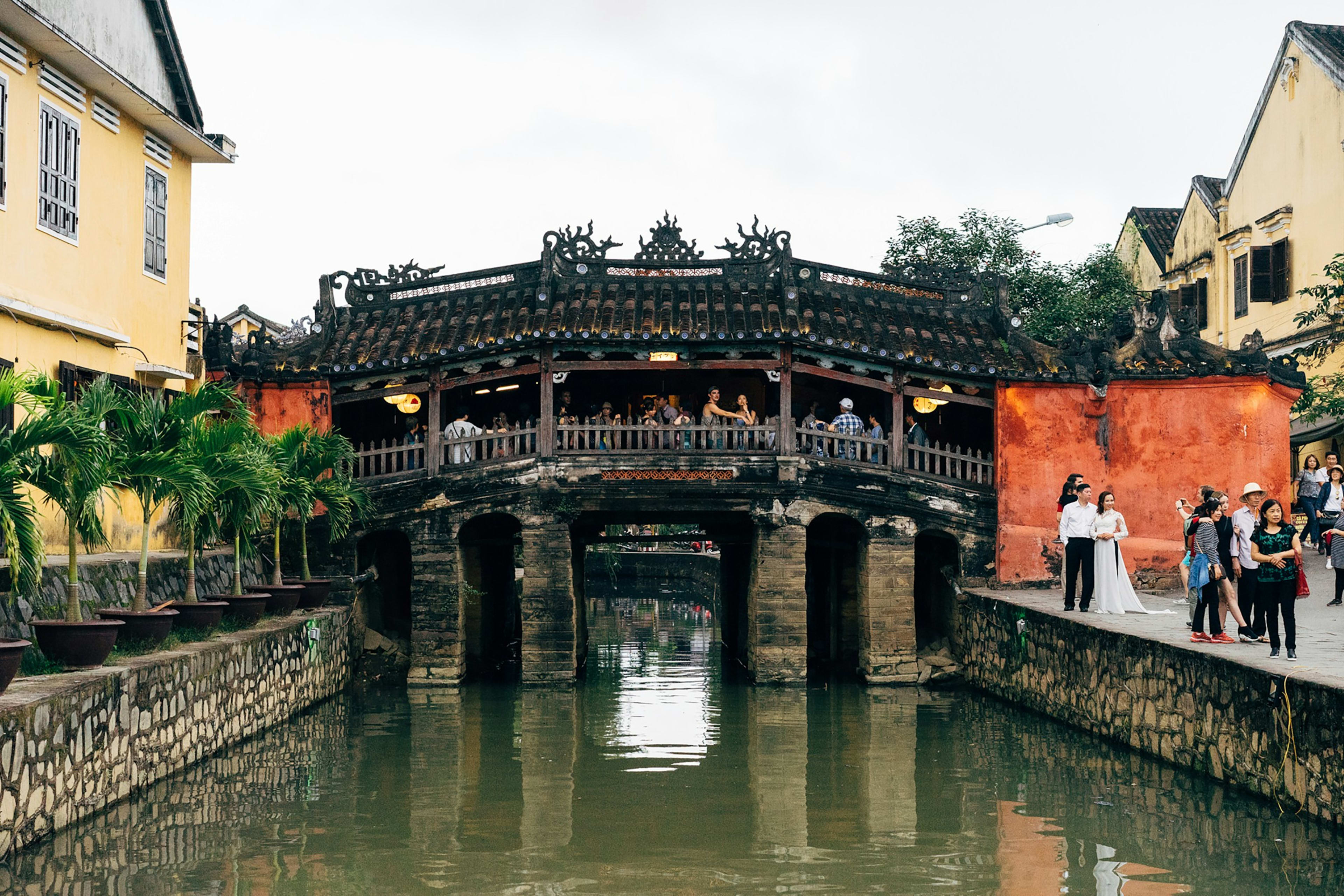 The Japanese Covered Bridge arching over a narrow canal in Hội An, its tiled roof and carved gable beside an ochre wall where visitors stop to pose.