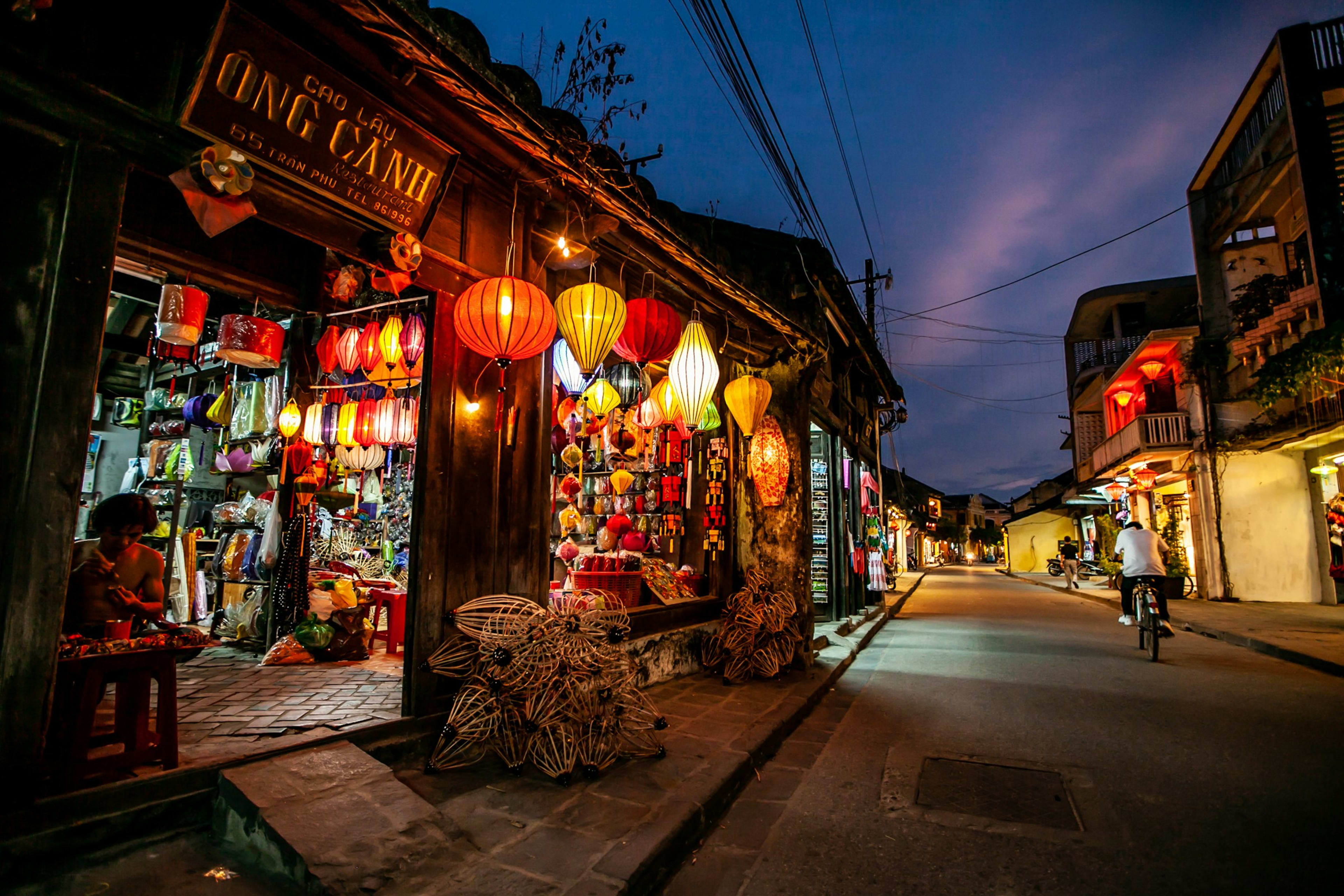 A Hội An shopfront hung with lit silk lanterns at dusk, the quiet street running past it and a cyclist in the distance.