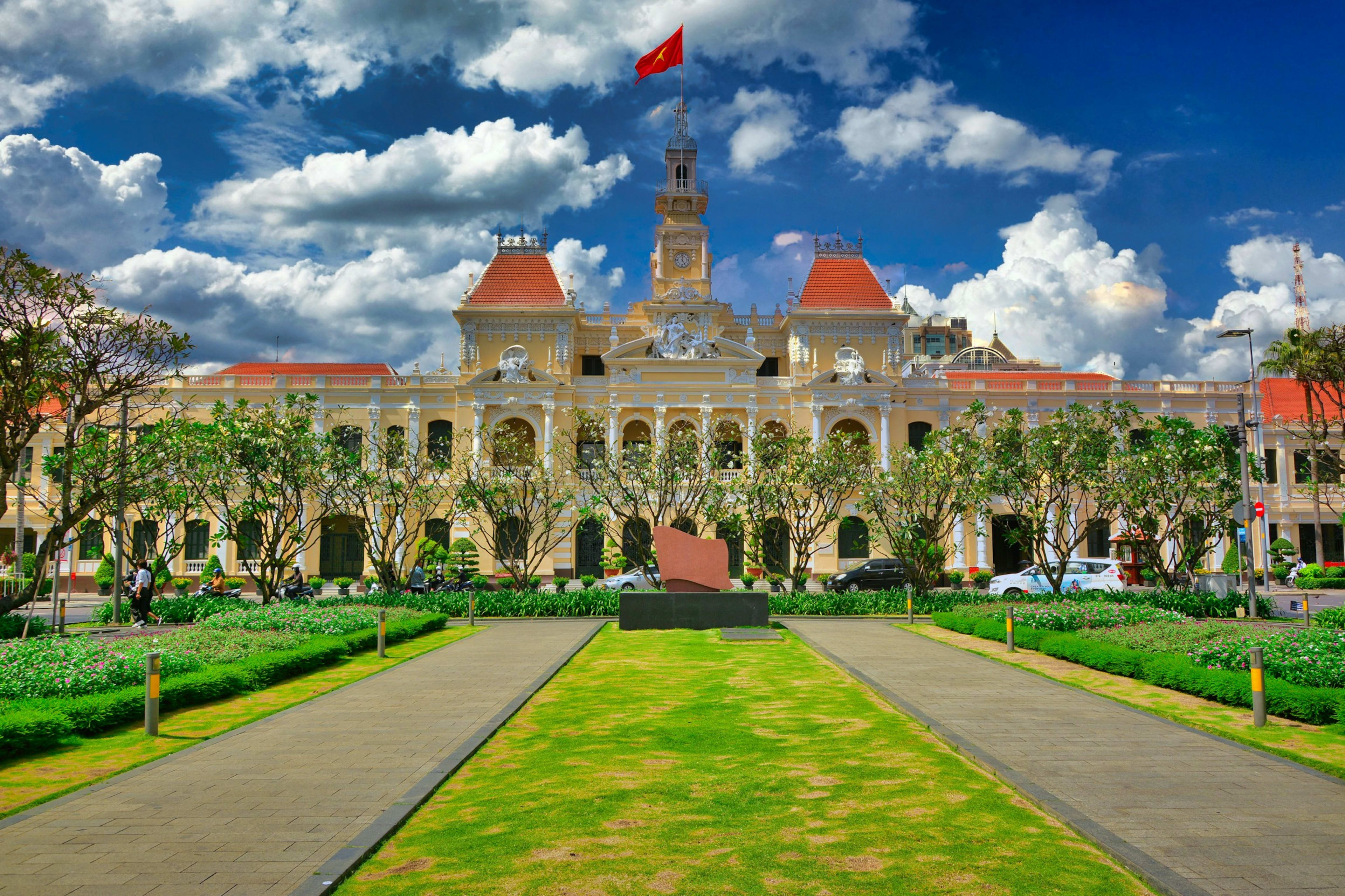 The People's Committee building's cream colonial façade and clock tower flying the Vietnamese flag, seen along the lawns of Nguyễn Huệ under piled cumulus.