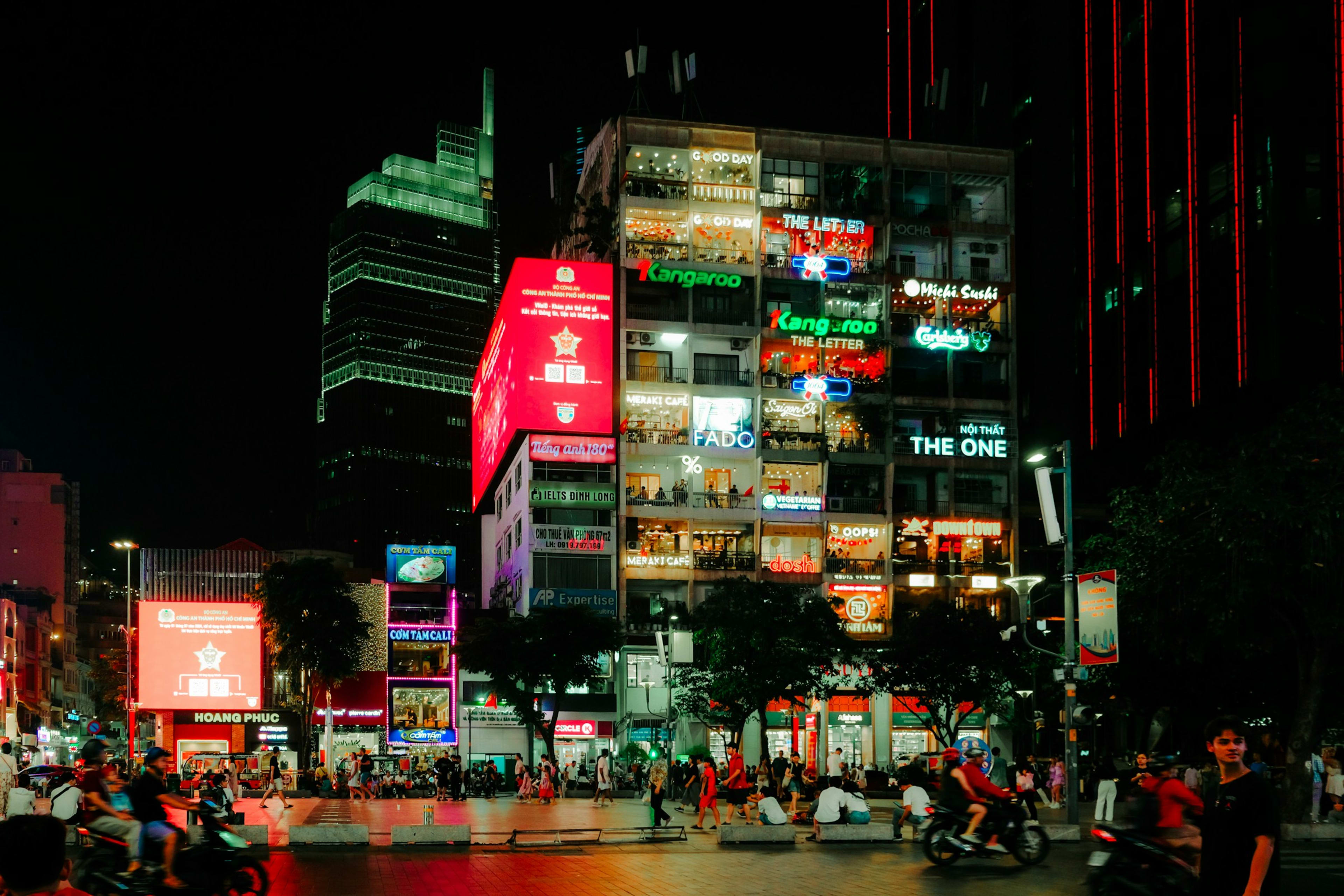 A Sài Gòn café apartment block lit up at night, every balcony a different neon sign, with motorbikes and crowds on the street below.