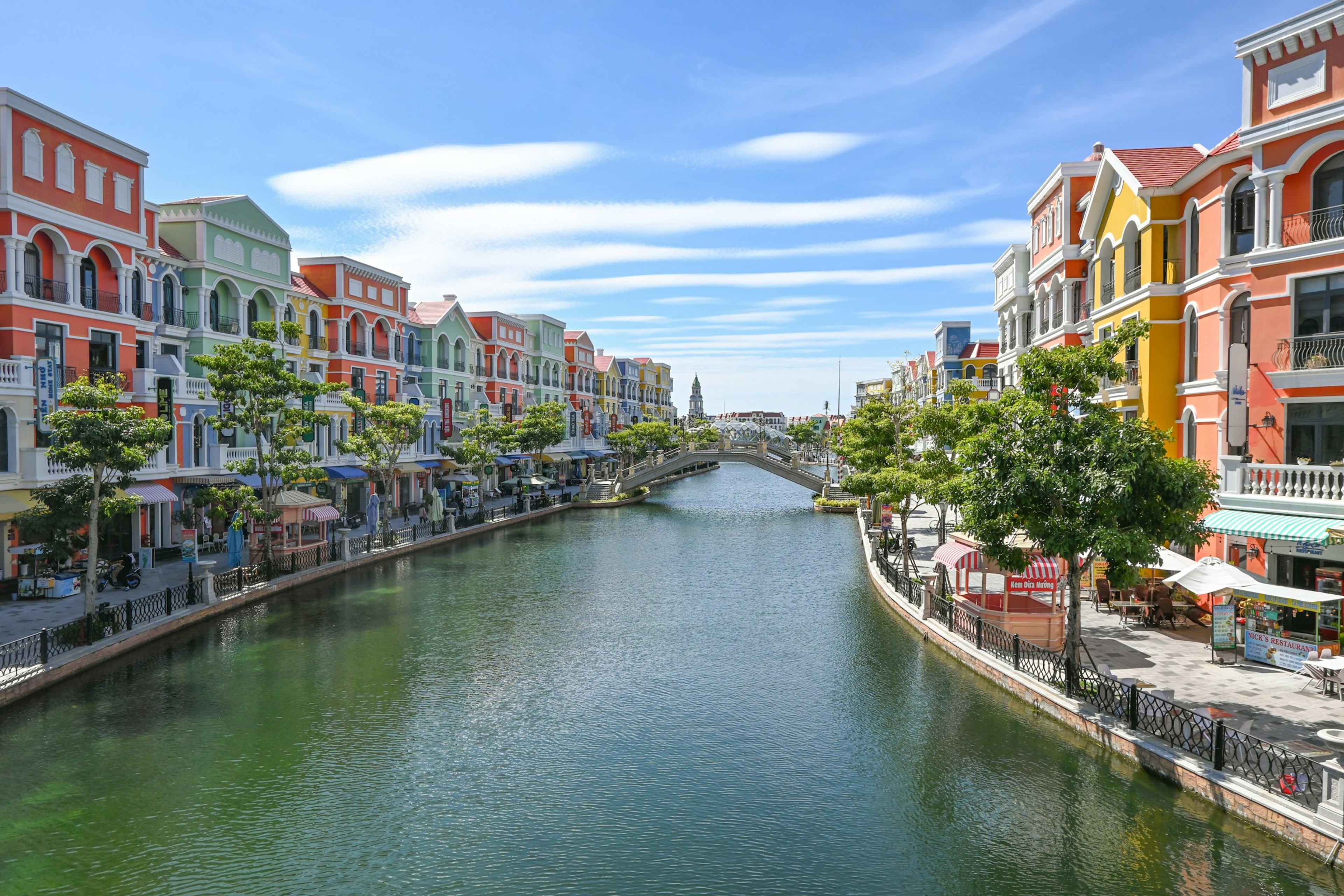 The same pastel canal seen from a bridge, colourful buildings down both banks with young trees and café umbrellas along the water.