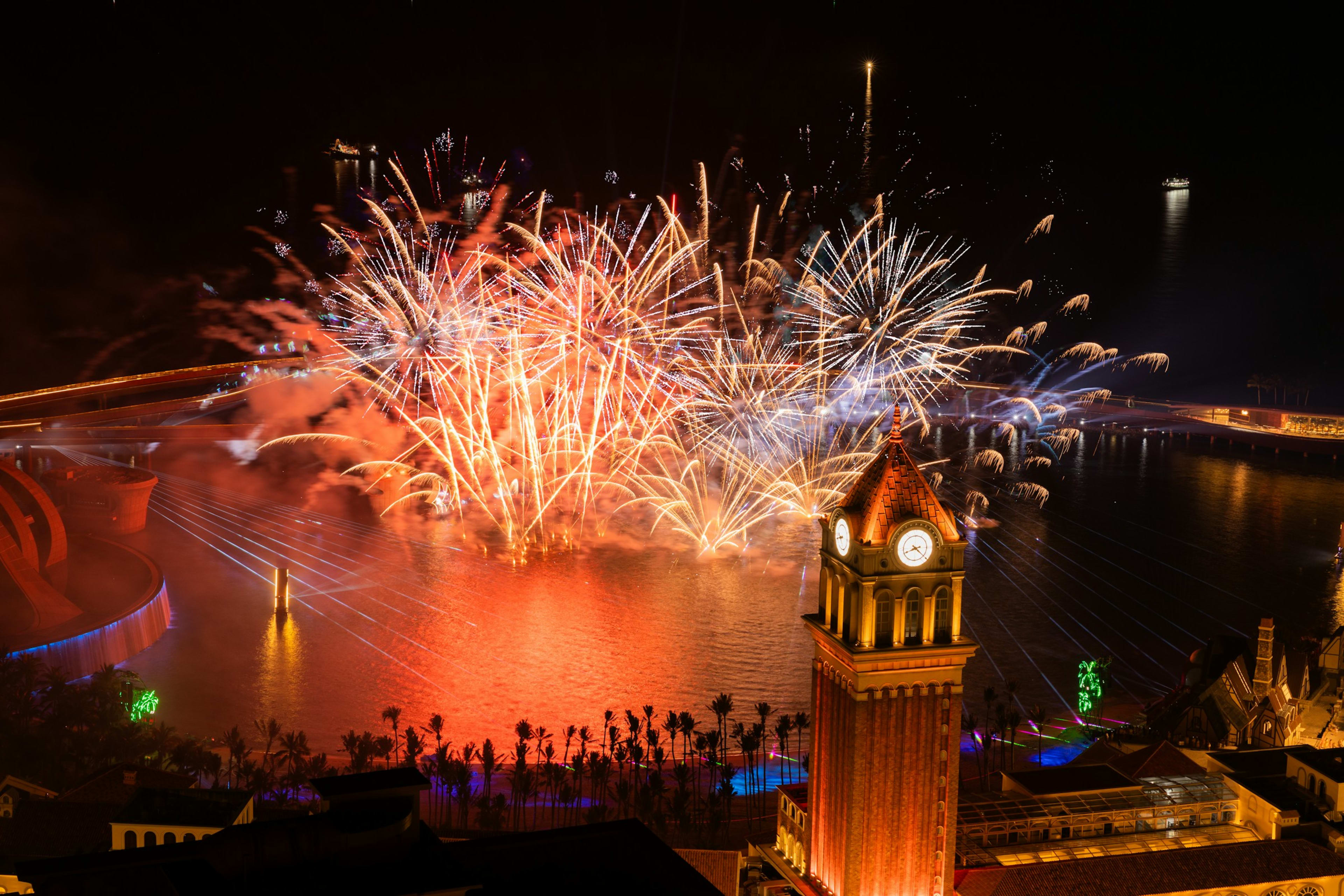 Fireworks bursting over the water beside a lit clock tower, palms in silhouette along the shore below.