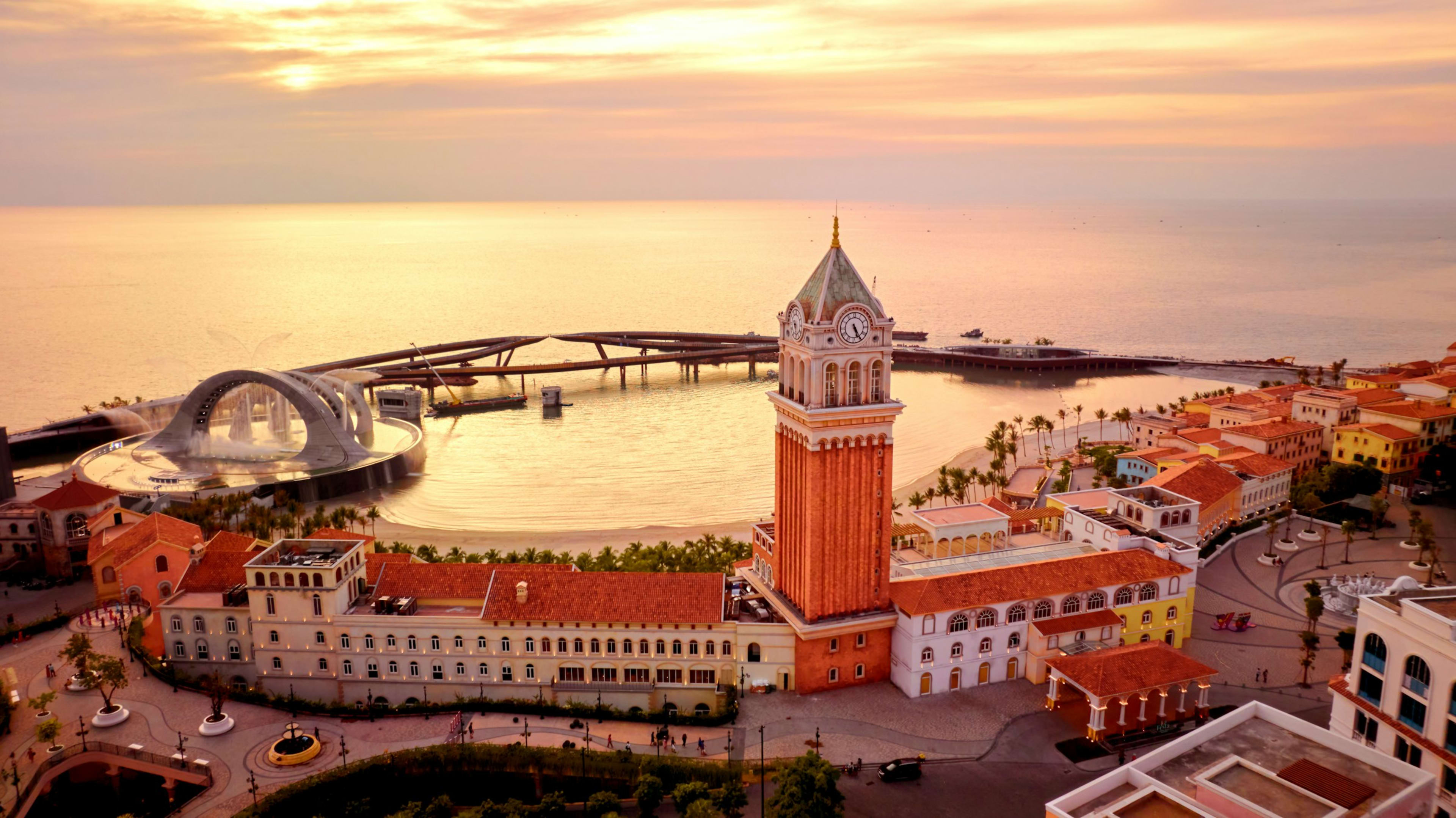 A resort quarter at sunset gathered around an orange clock tower, a curved footbridge reaching out over a calm gold sea.