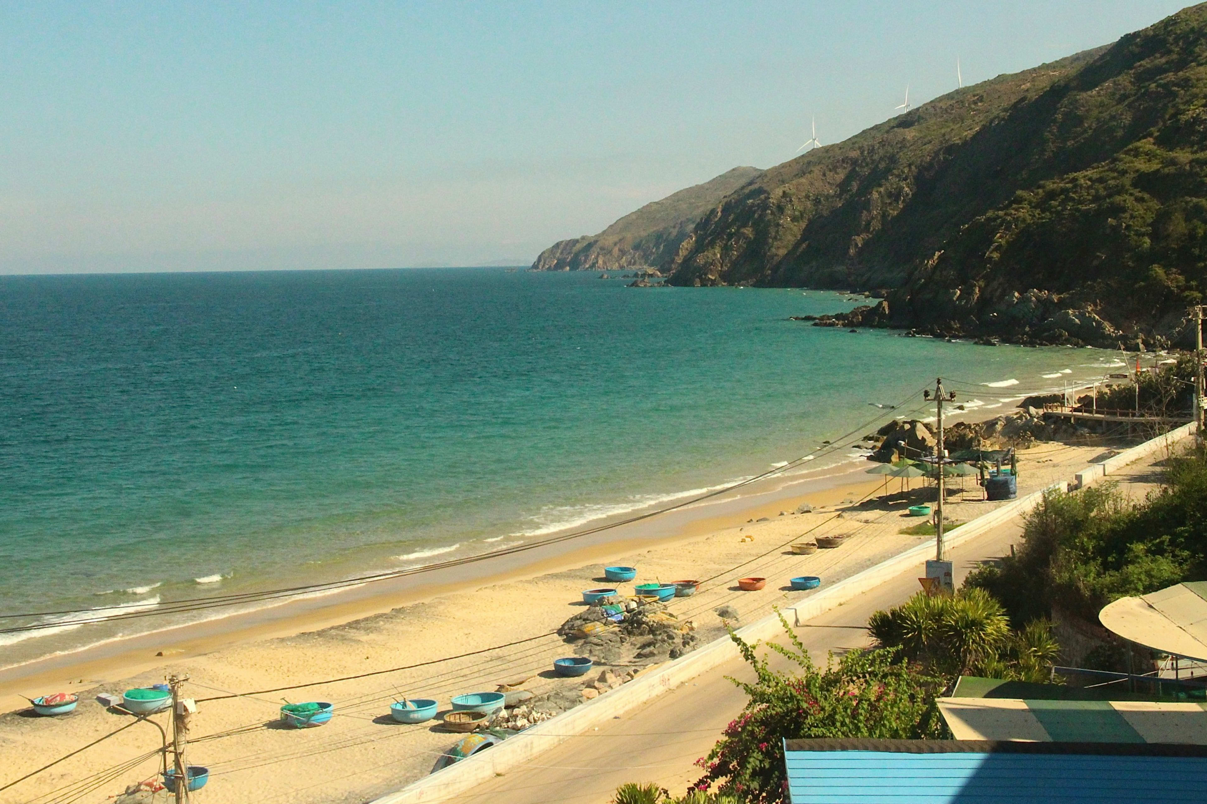 A long crescent beach with basket boats drawn up on the sand, a mountain headland behind and wind turbines along its ridge.