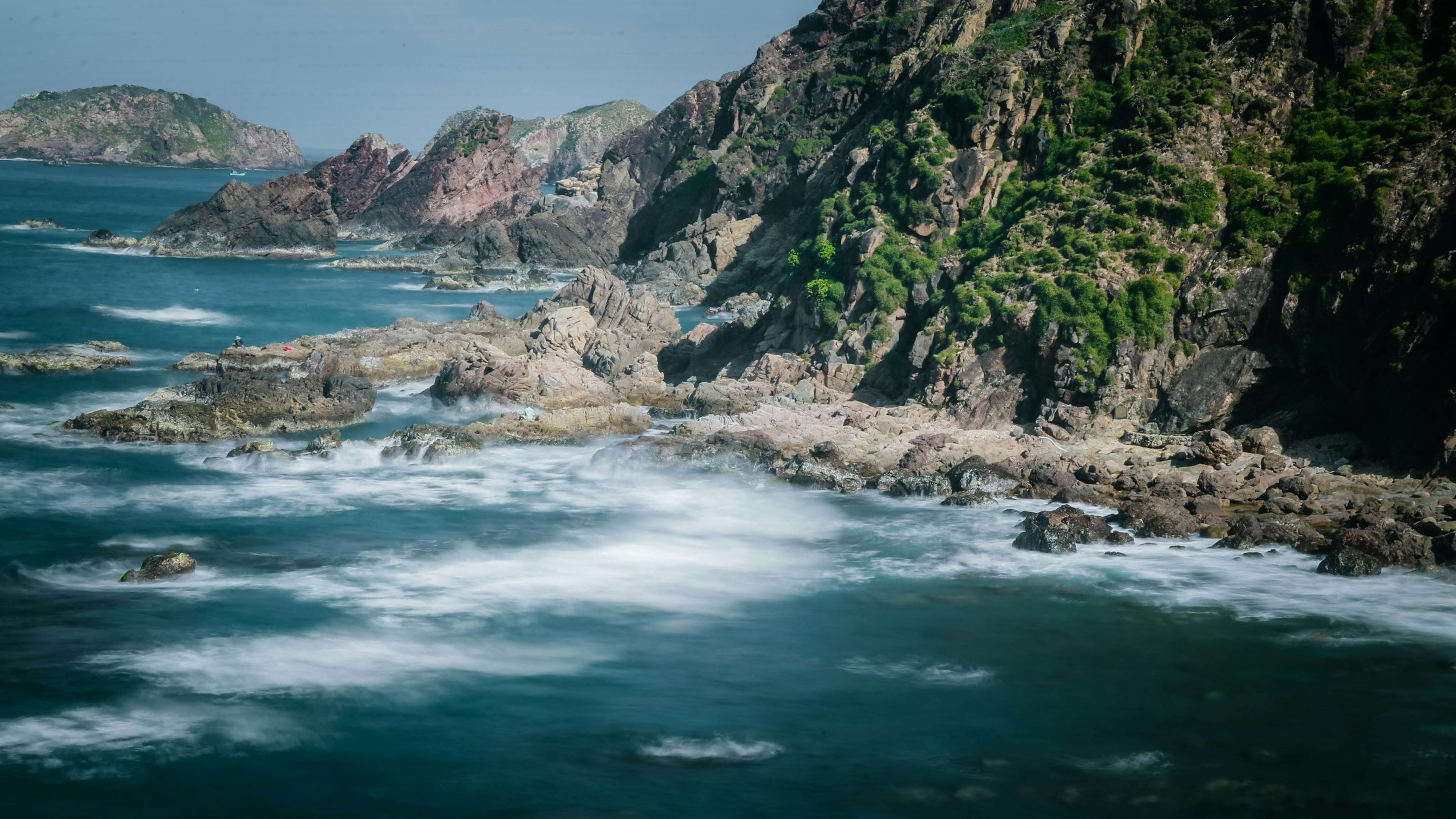 Waves washing over rocks below a steep headland, a long exposure blurring the surf to white mist.