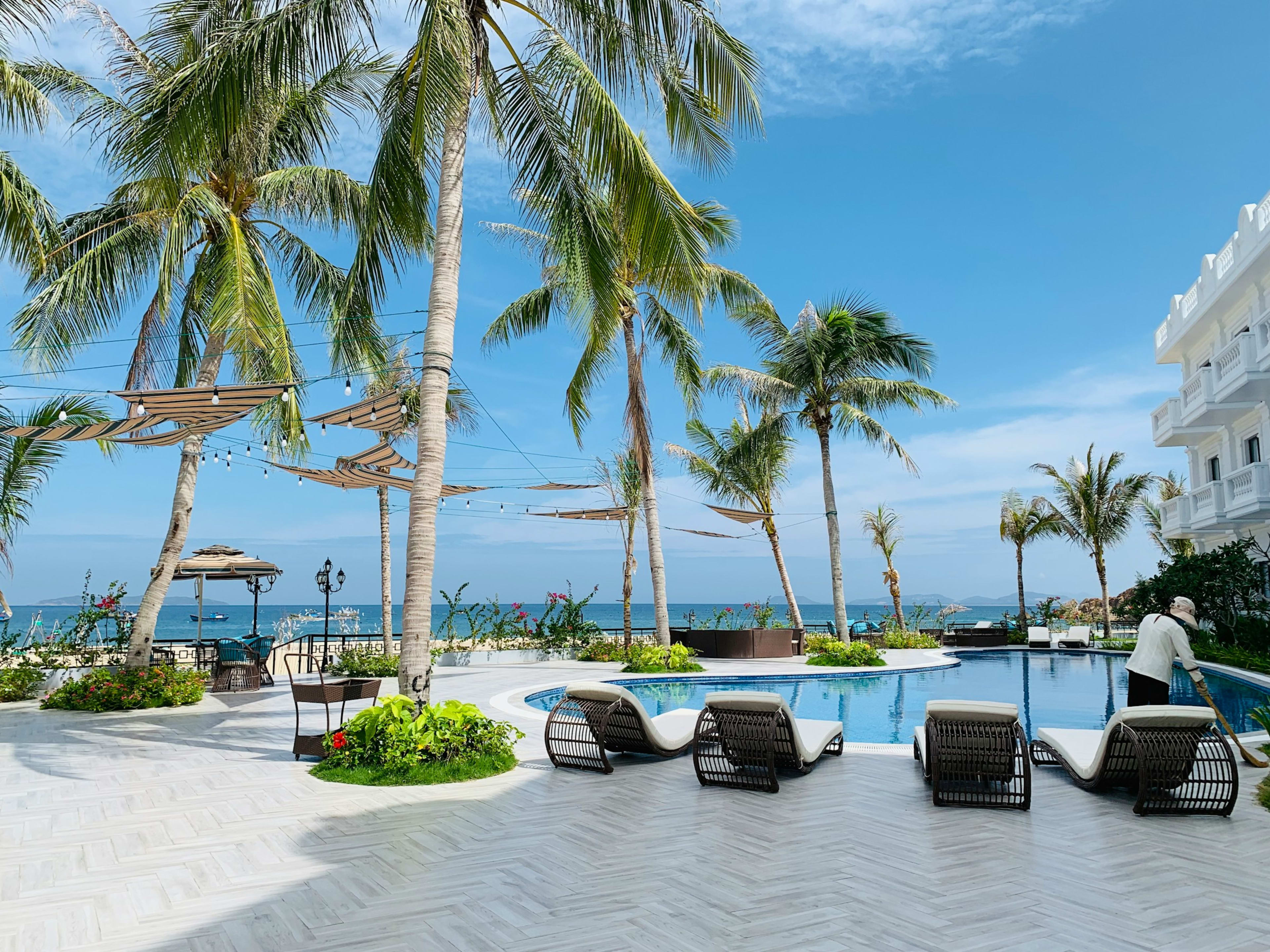 Sun-loungers beside a pool ringed with coconut palms and shade sails, the open sea just beyond the terrace.