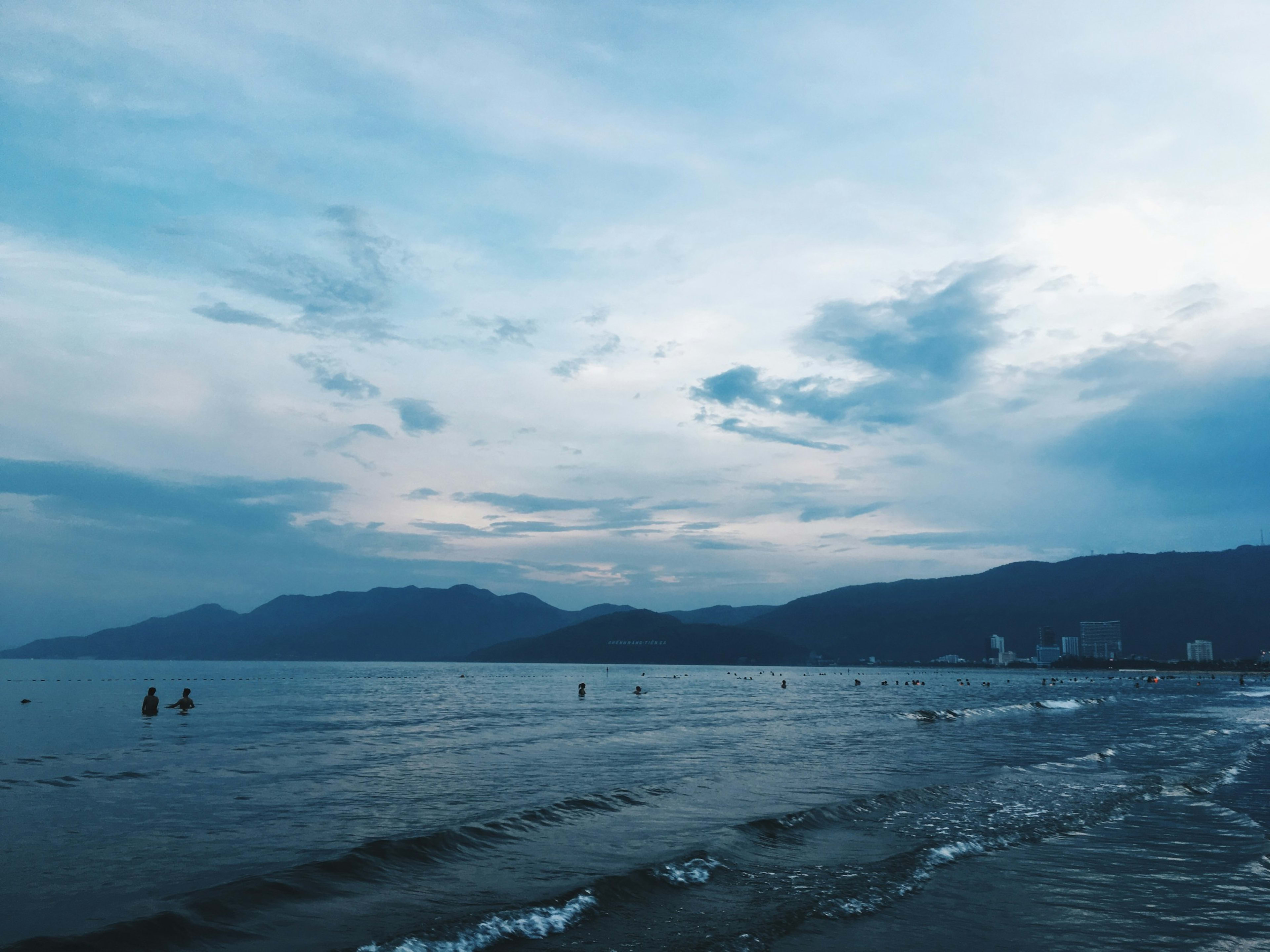 A grey-blue evening at the beach with swimmers in the shallows, mountains along the far shore and town buildings at the end of the bay.