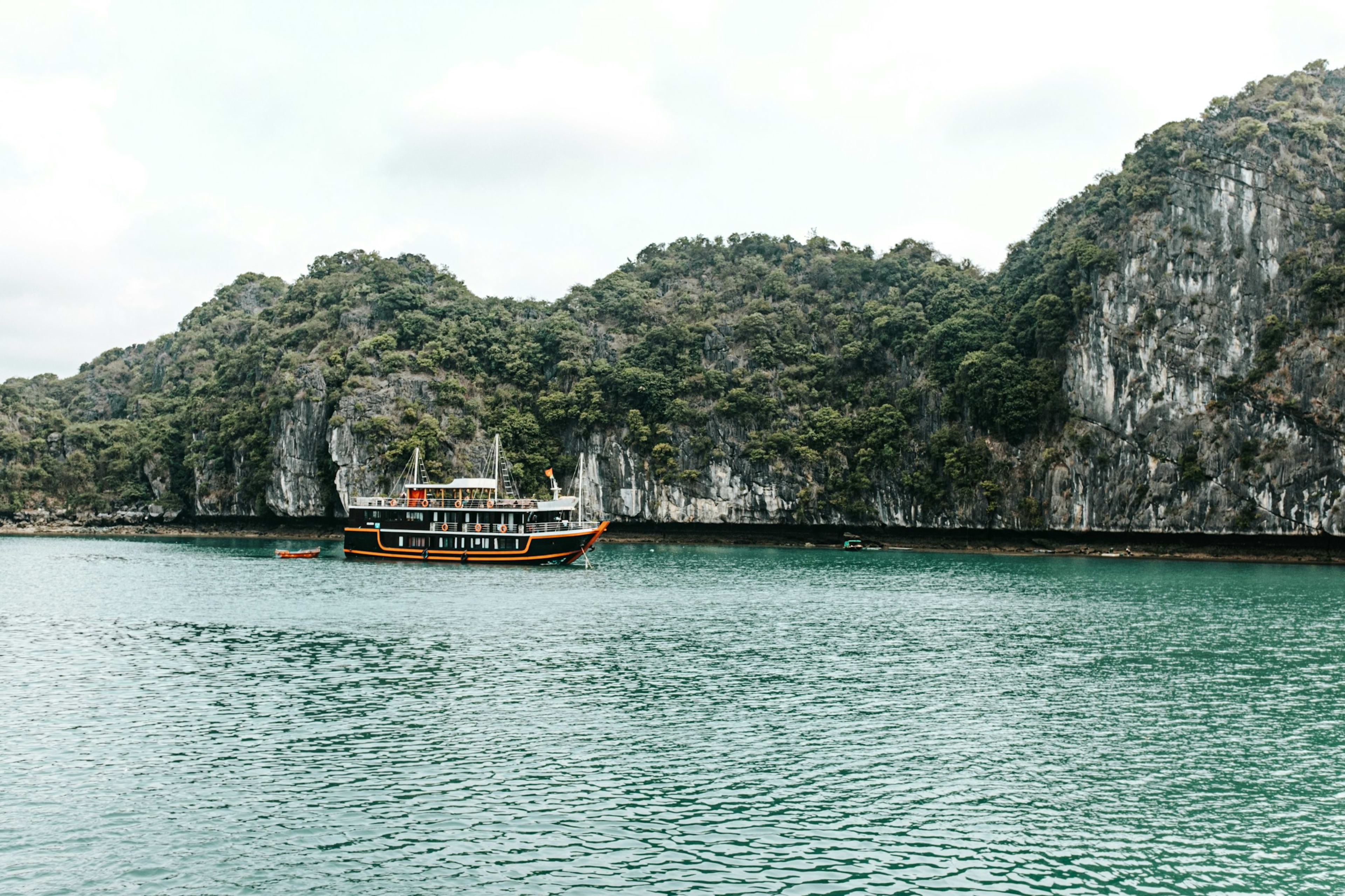 A wooden cruise junk anchored close under a wall of forested limestone cliffs in flat green water.