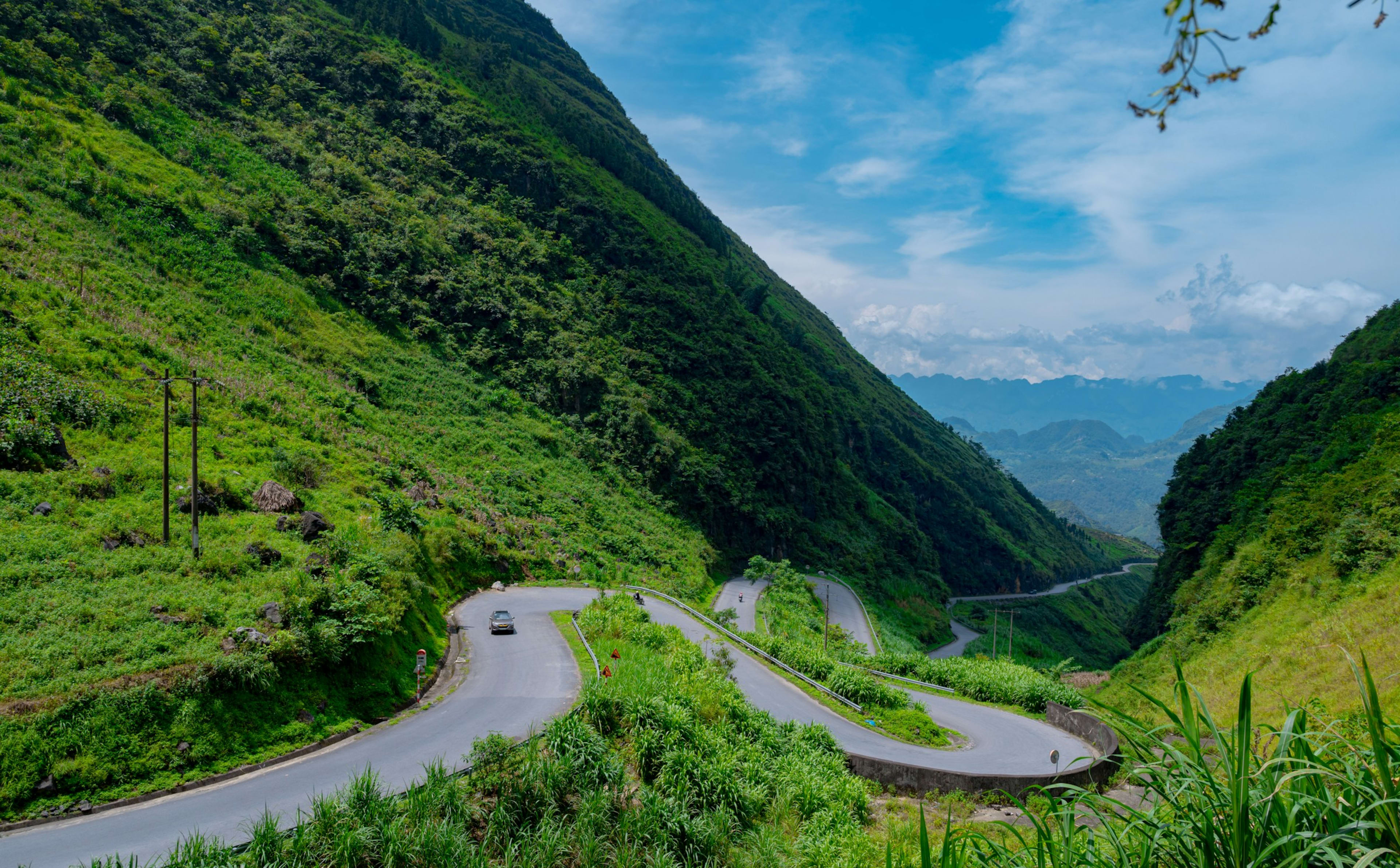 A mountain road switchbacking down a steep green valley, a single car on one of the bends and ridges receding into haze.