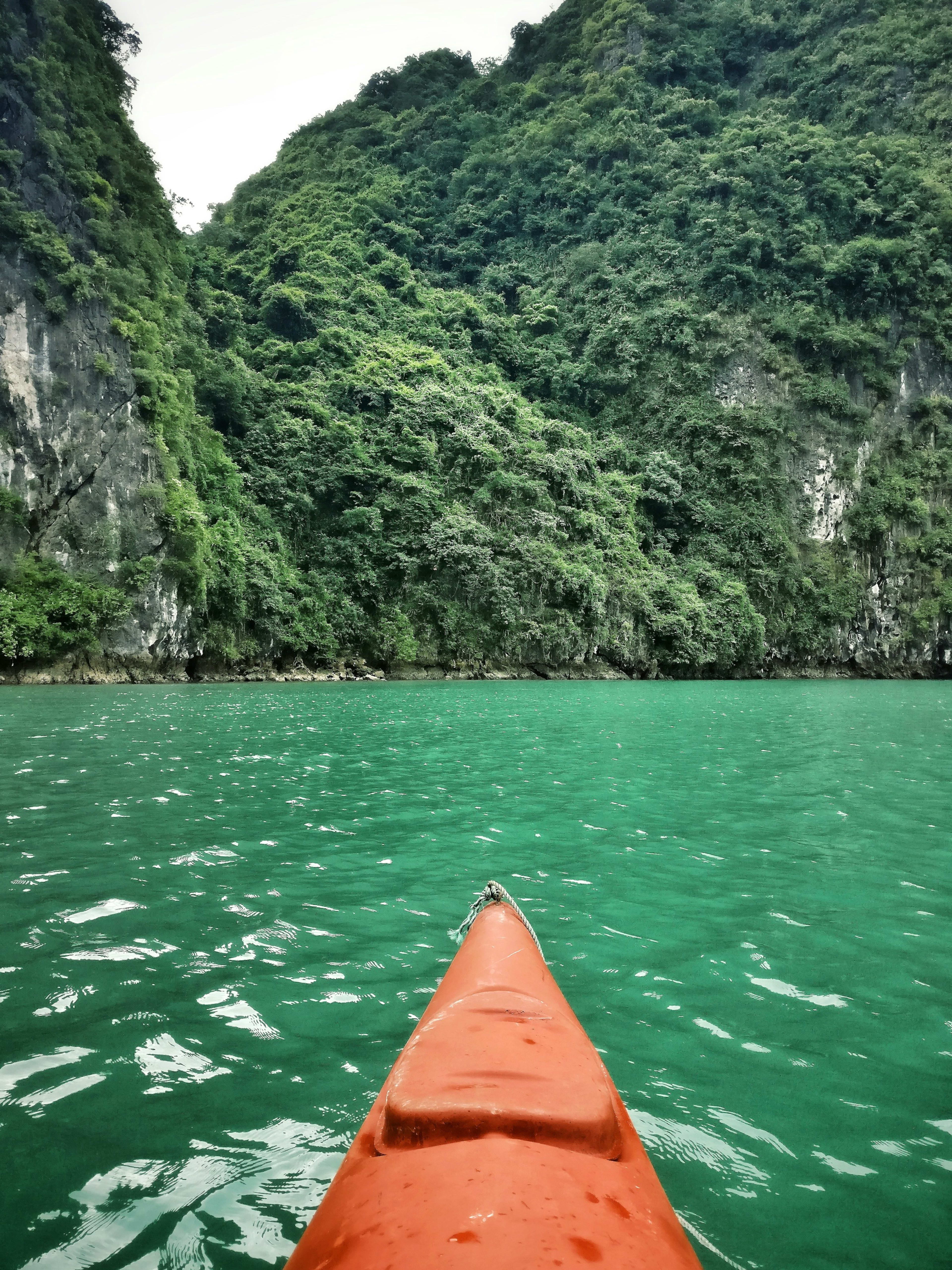 The orange bow of a kayak on flat emerald water in front of a limestone cliff furred with jungle.