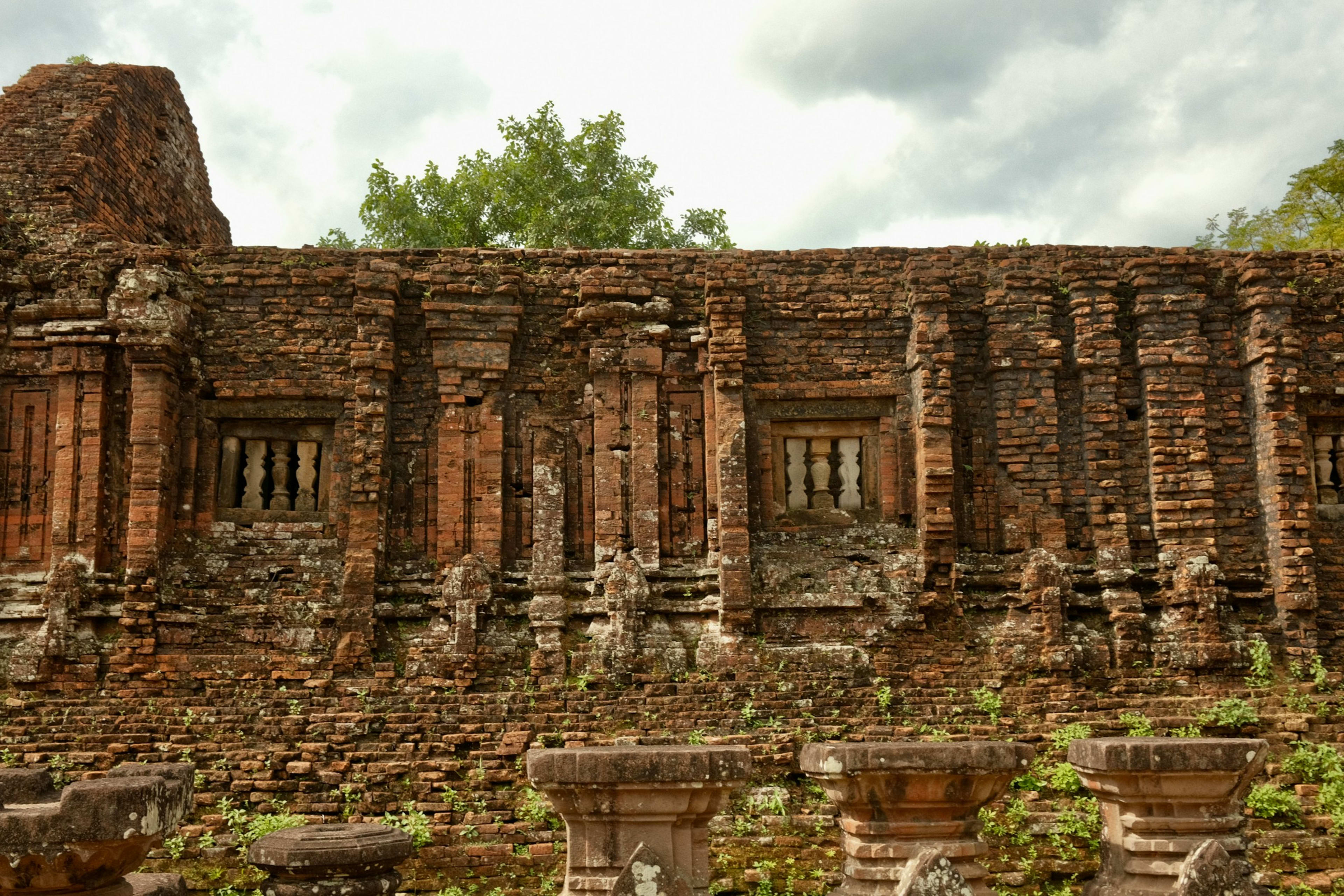 A long weathered brick wall of a Chăm temple with carved pilasters and lattice windows, fallen columns lying in front of it.