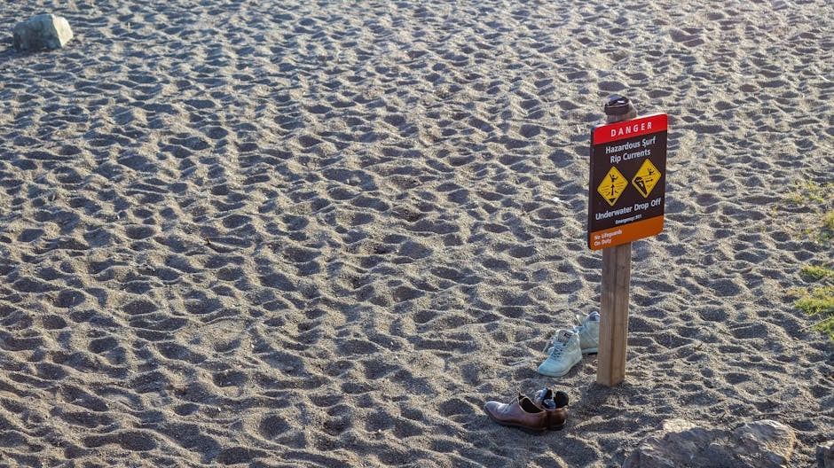 A beach warning sign stands on the sandy shore with two pairs of shoes nearby, emphasizing caution.