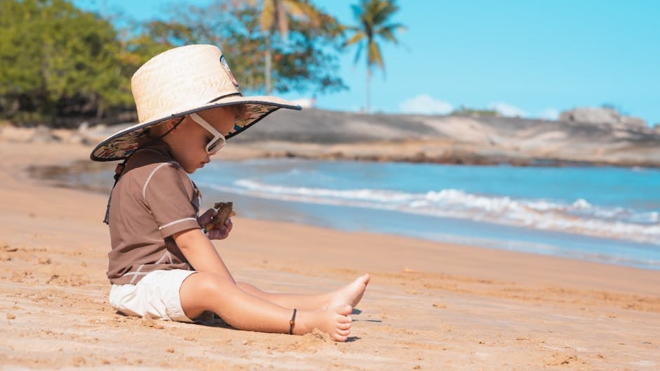 A young child wearing a sunhat sits on a sandy tropical beach playing with a toy, enjoying a sunny day.