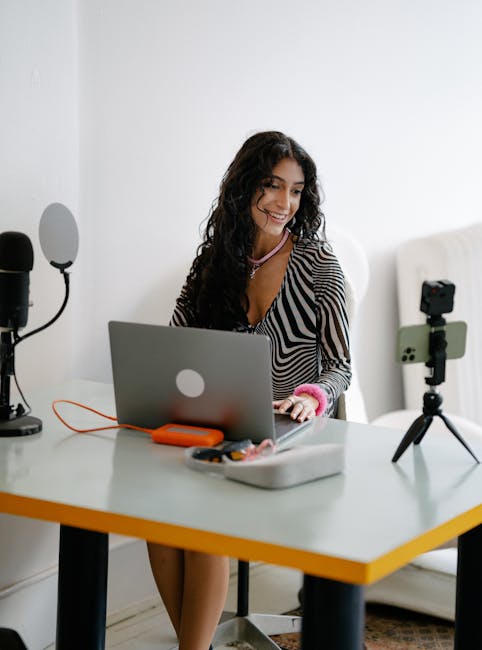 Smiling woman sits at desk using laptop, with camera and microphone setup for remote work.