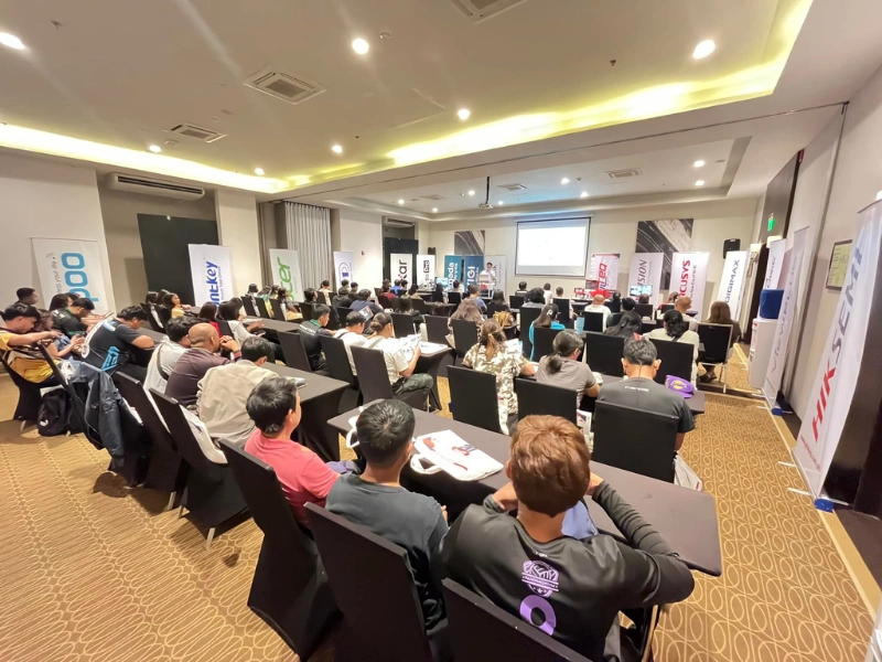 A seminar room filled with attendees listening to a presentation, with brand banners such as Ducky, Hiksemi, and Rapoo displayed around the room.