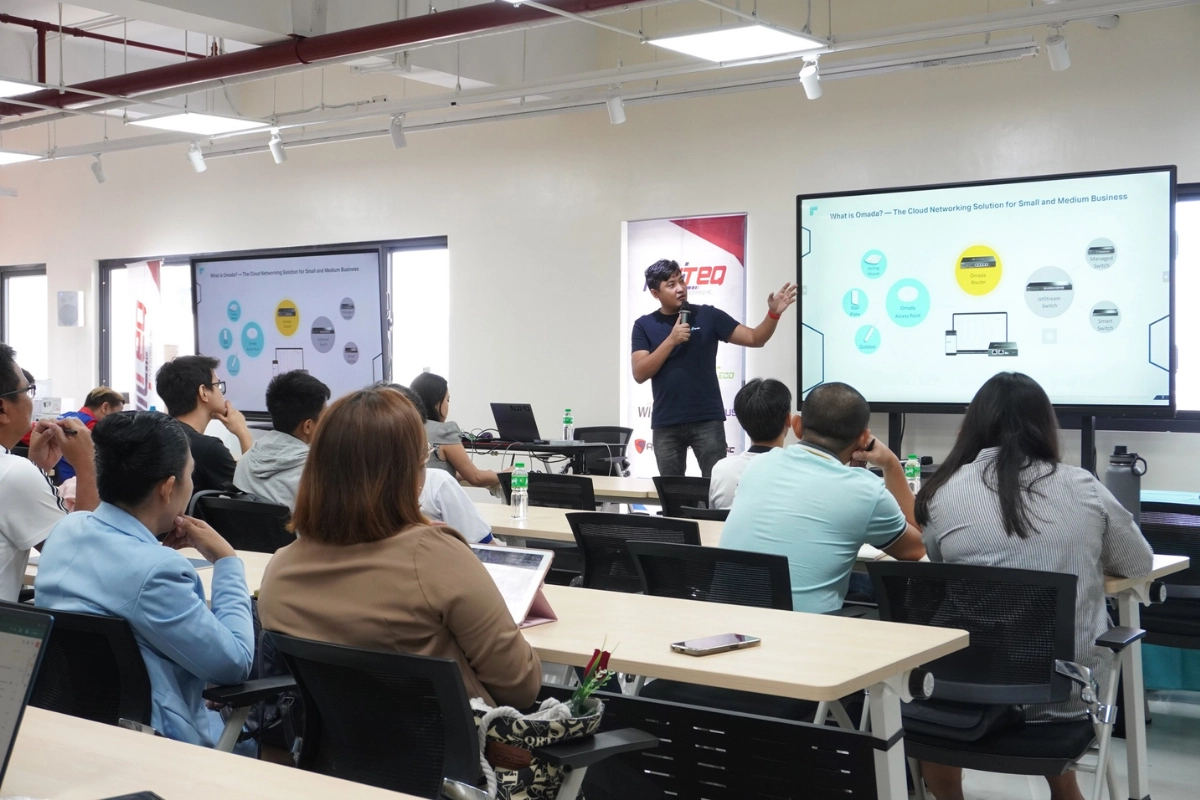 A speaker presents to an attentive audience in a modern classroom, pointing to a screen displaying a digital presentation. The atmosphere is focused and engaging.