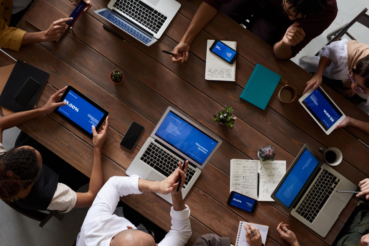 Overhead view of a diverse business team collaborating around a wooden table, using laptops, tablets, and smartphones to view data and discuss strategies.