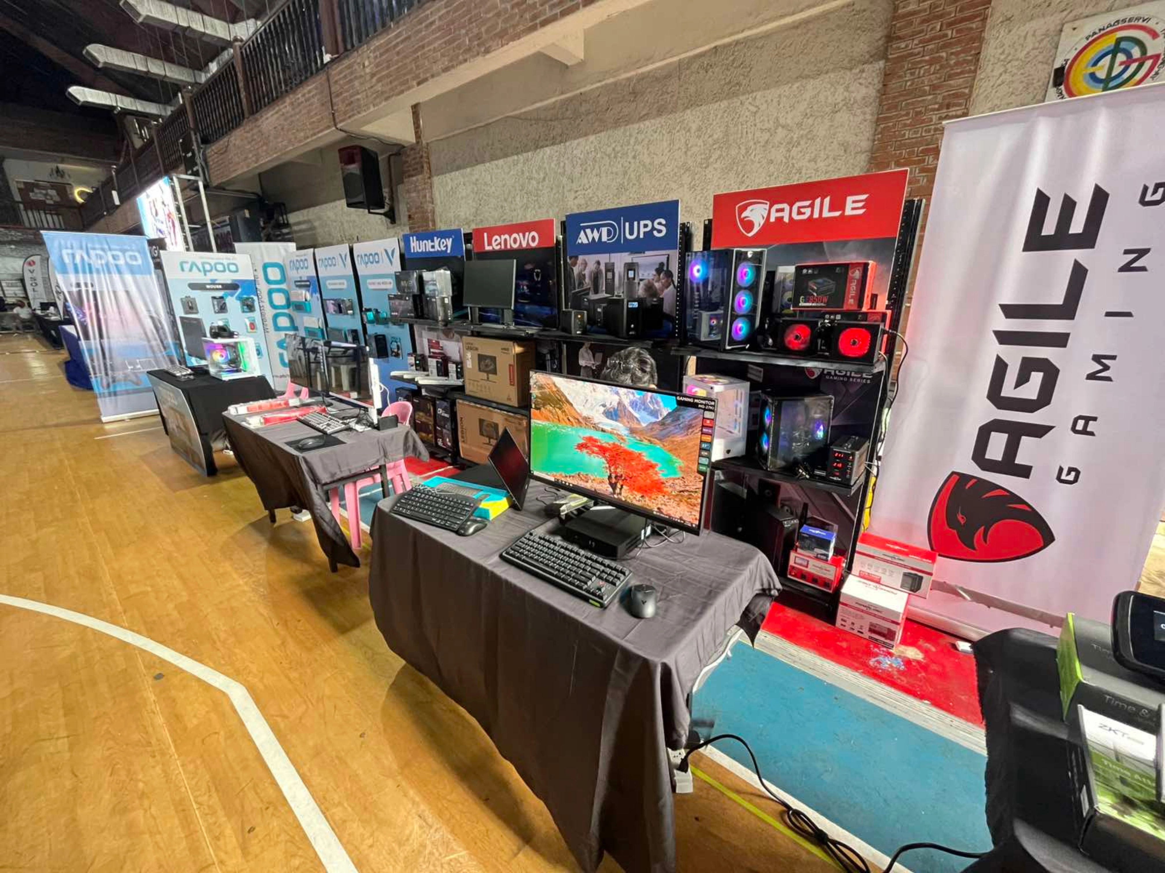 Trade show booth displaying gaming PCs, monitors, keyboards, and other IT products from brands like Agile Gaming, Lenovo, and AMD UPS.