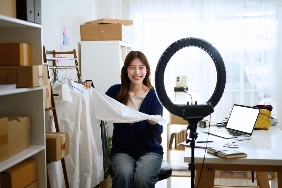 Smiling young woman preparing to live stream or record an e-commerce video, holding a white shirt next to a ring light, smartphone, and laptop in a home office with shipping boxes.