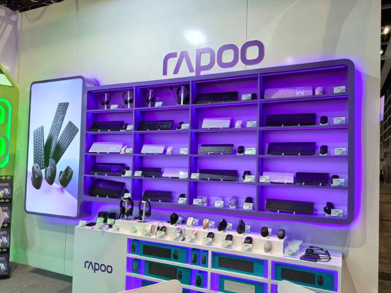 A Rapoo booth display featuring shelves of keyboards, mice, and headsets illuminated by purple lighting.