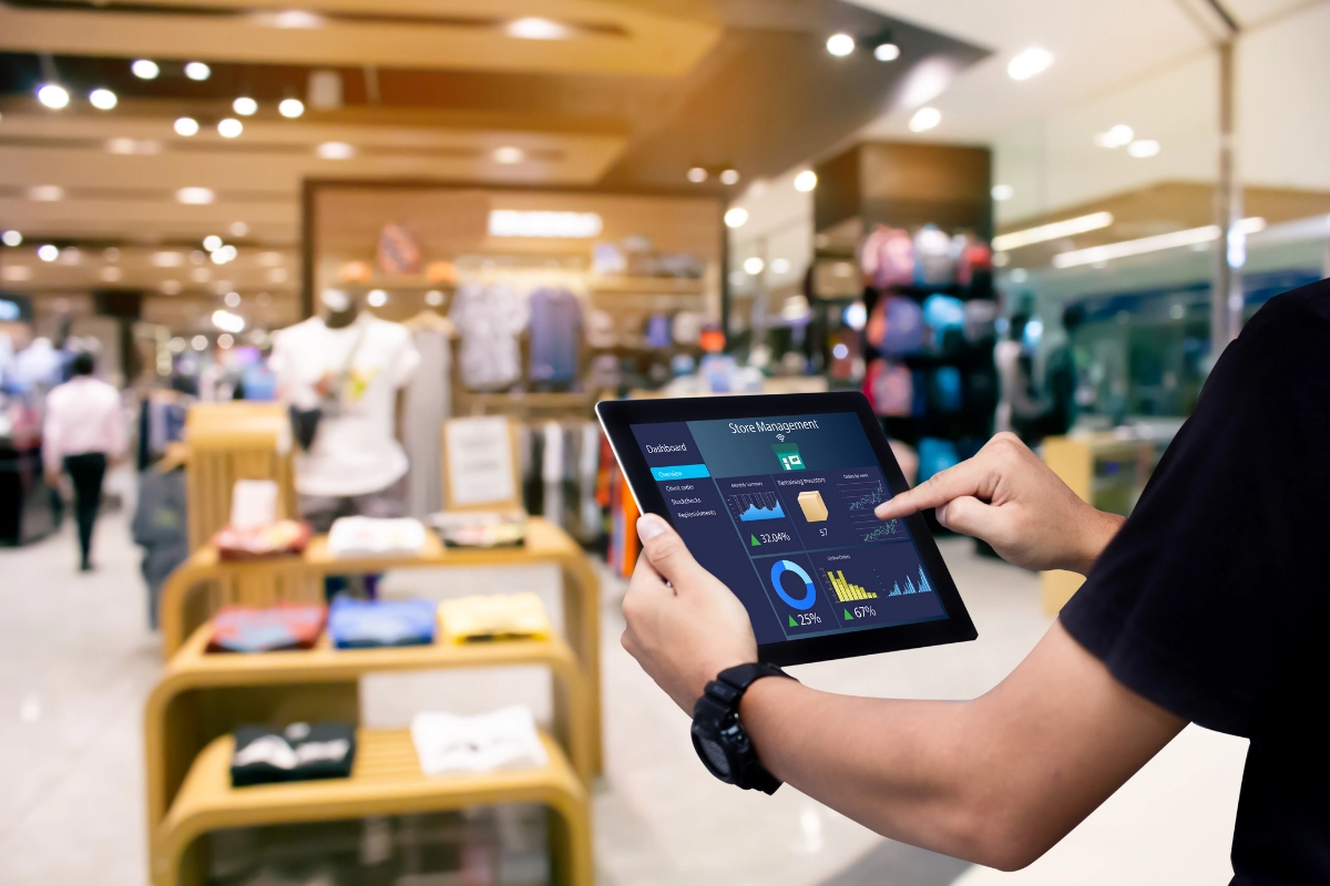 Person using a tablet to view a "Store Management" dashboard with retail analytics, pie charts, and sales data, with a blurred apparel store interior in the background.