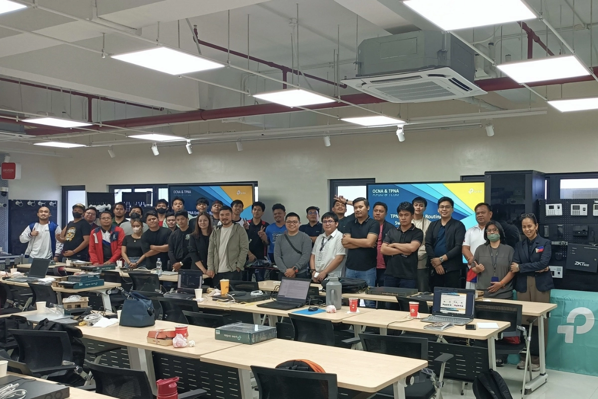 Large group of professionals and trainees posing for a group photo in a modern classroom setting with desks, laptops, and a presentation screen displaying "TP-Link DCNA & TPNA" certification course information.