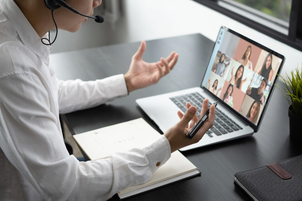Person in a white shirt on a video call, gesturing with hands, wearing a headset. Open laptop shows multiple participants in a virtual meeting.
