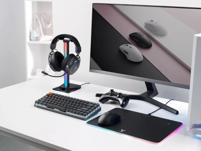 A modern computer setup featuring a mechanical keyboard, gaming mouse, headset on a stand, and monitor—all with RGB lighting accents.