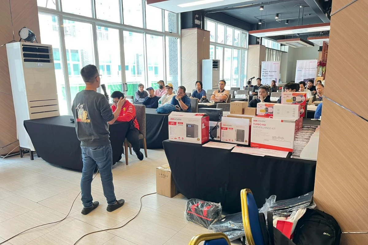 A man presents to an engaged audience in a bright room, with surveillance kit boxes displayed on tables. The atmosphere is attentive and professional.