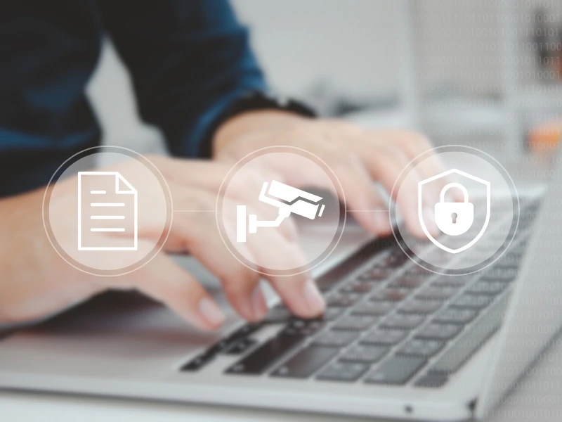 Hands typing on a laptop keyboard with overlay icons of a document, surveillance camera, and padlock, symbolizing digital security and privacy.