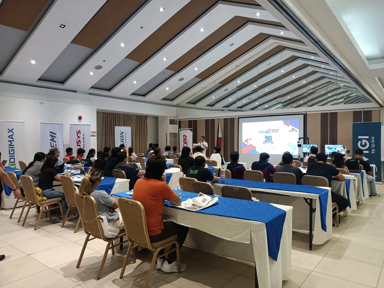 People attending a presentation in a modern conference room. Branded banners line the walls, and a speaker stands near a large projection screen.