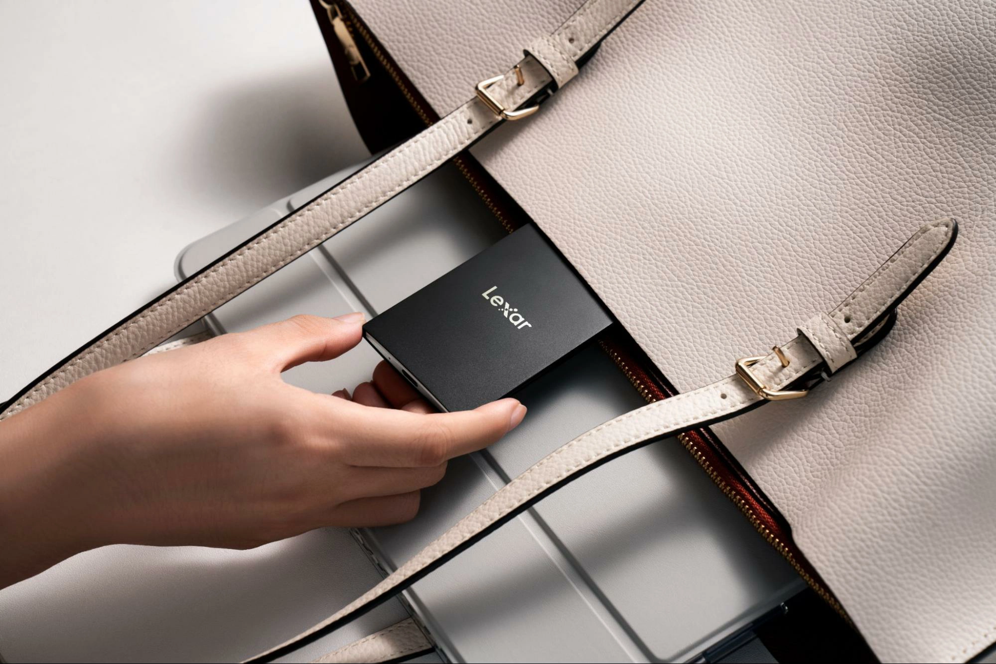 A hand placing a compact, black Lexar external SSD into a cream-colored handbag, emphasizing portability and size.