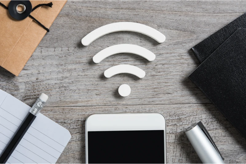 Flat lay of a wooden desk displaying a smartphone, WiFi symbol made of white shapes, notebook, pen, and envelope, suggesting connectivity and communication.