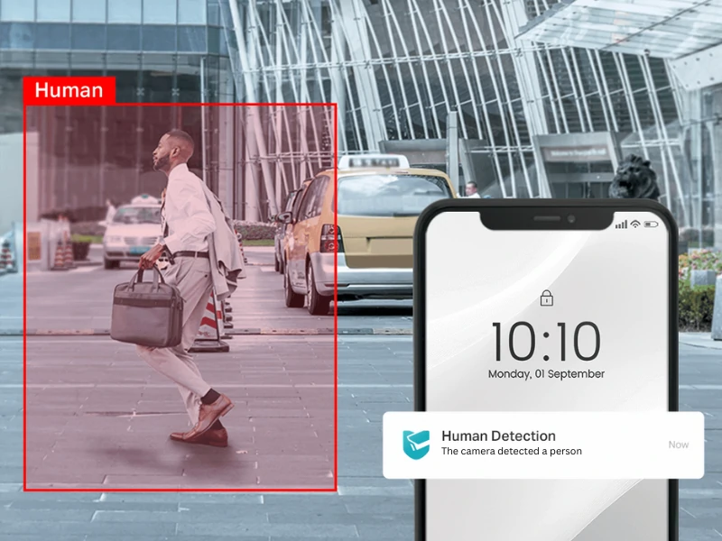 A man in a light suit with a briefcase walks briskly past yellow taxis in a city. A smartphone shows an alert for "Human Detection."
