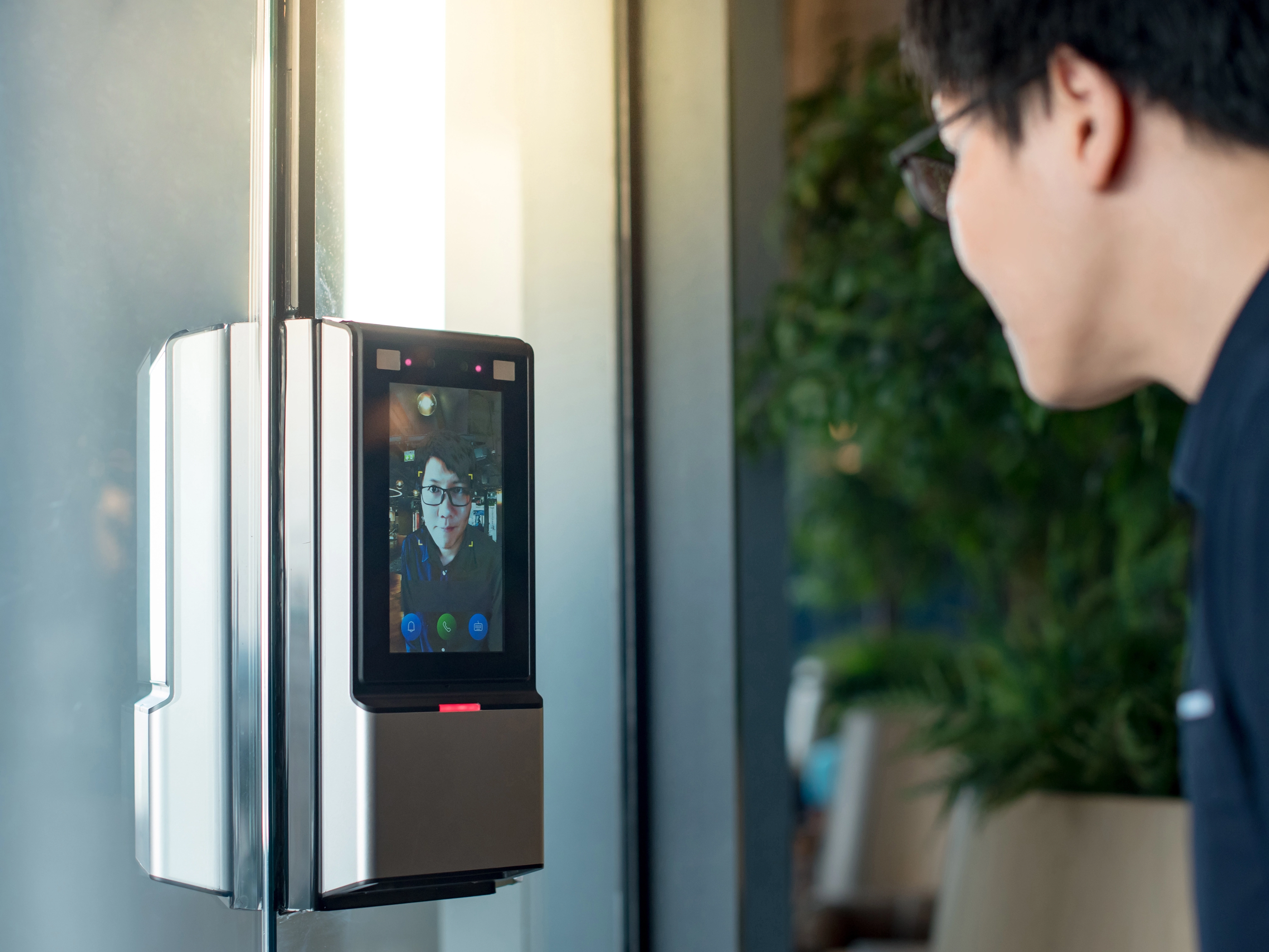A person facing a facial recognition device on a glass wall. The screen displays the individual's image with a green checkmark, indicating successful access.