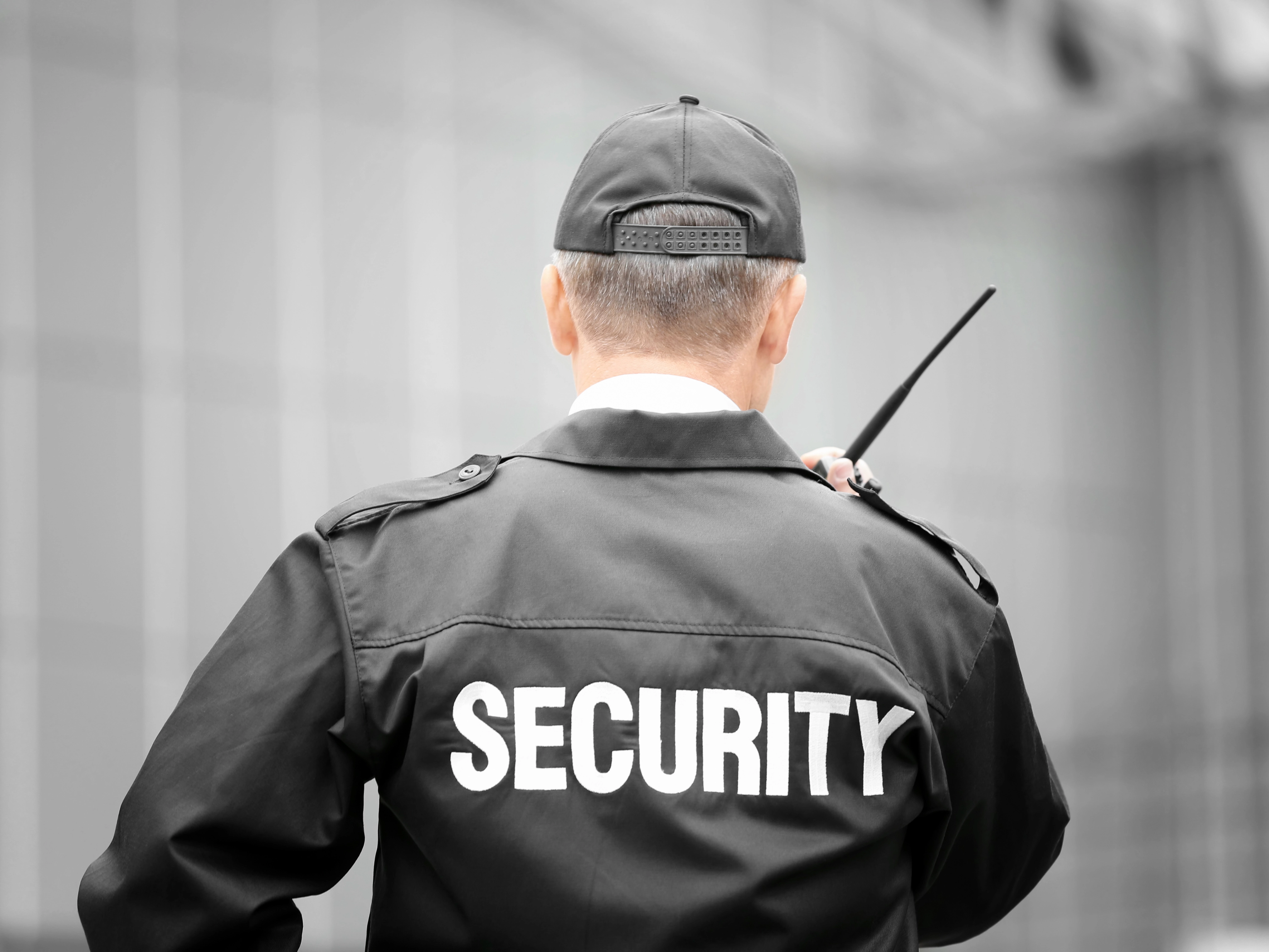 A security guard in uniform and cap stands with his back facing the camera, holding a walkie-talkie. The background is a blurred building facade.