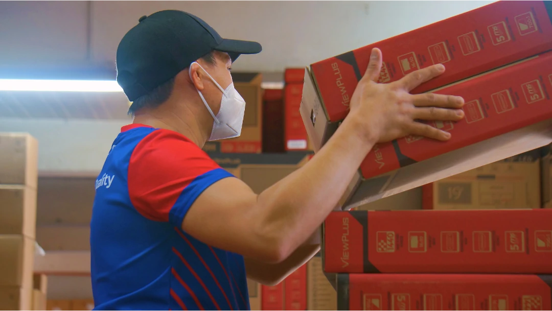 Warehouse worker in a blue and red uniform, wearing a mask and cap, carefully stacking red boxes labeled "VIEWPLUS." Shelves filled with supplies.