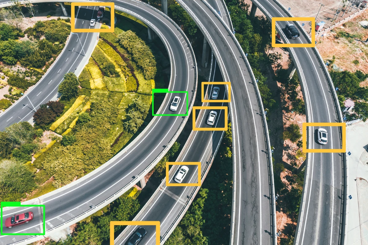 Aerial view of intersecting highways with multiple cars, each highlighted by orange or green boxes. Green areas with trees surround the roads.