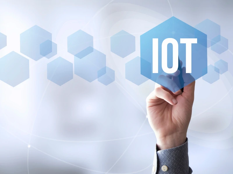 A hand touches a blue hexagon labeled "IOT" on a blurred background with interconnected hexagons, symbolizing technology and connectivity.