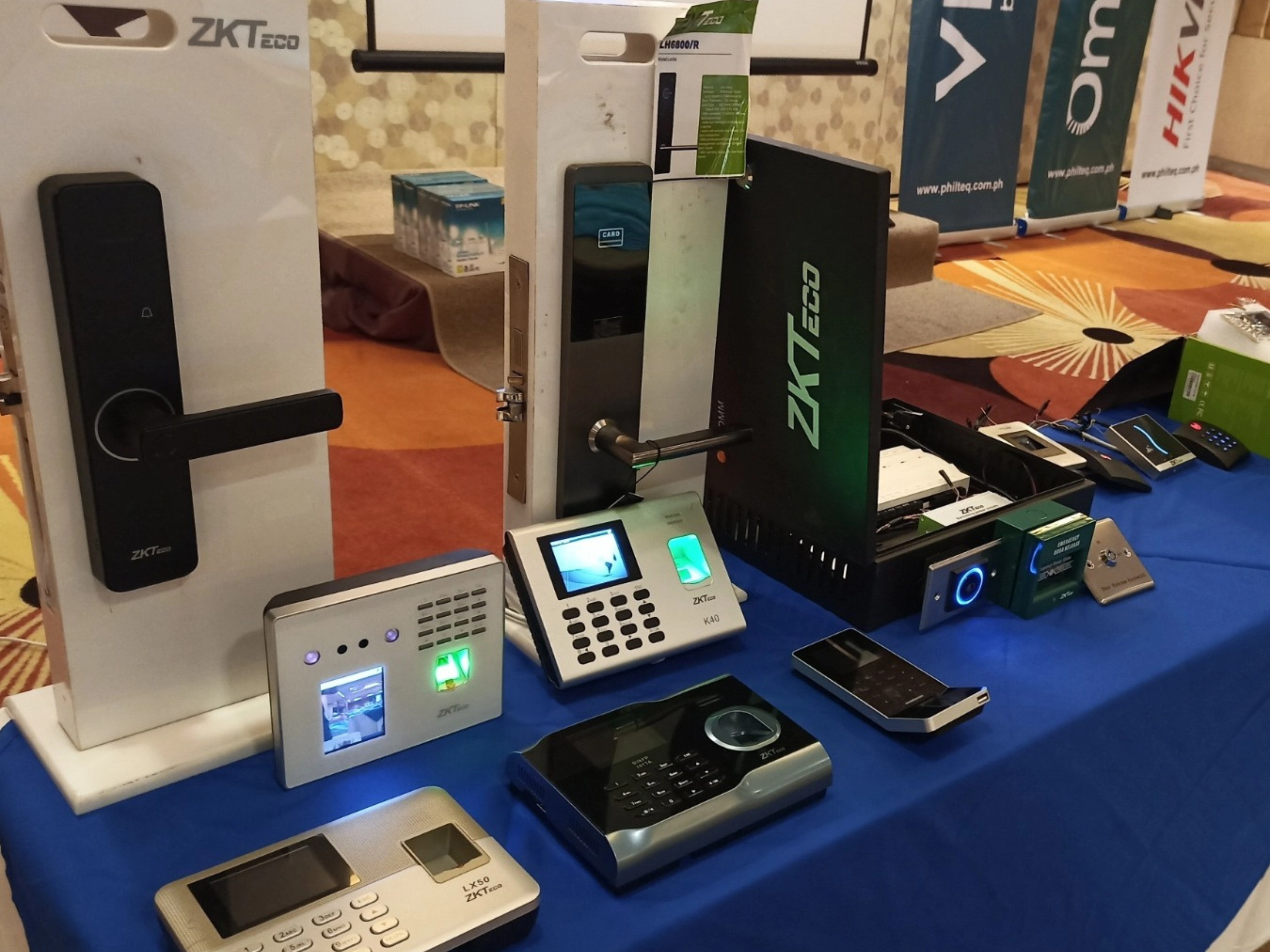 A table displays various ZKTeco security devices, including biometric scanners and digital locks, set on a blue cloth at a tech exhibition.
