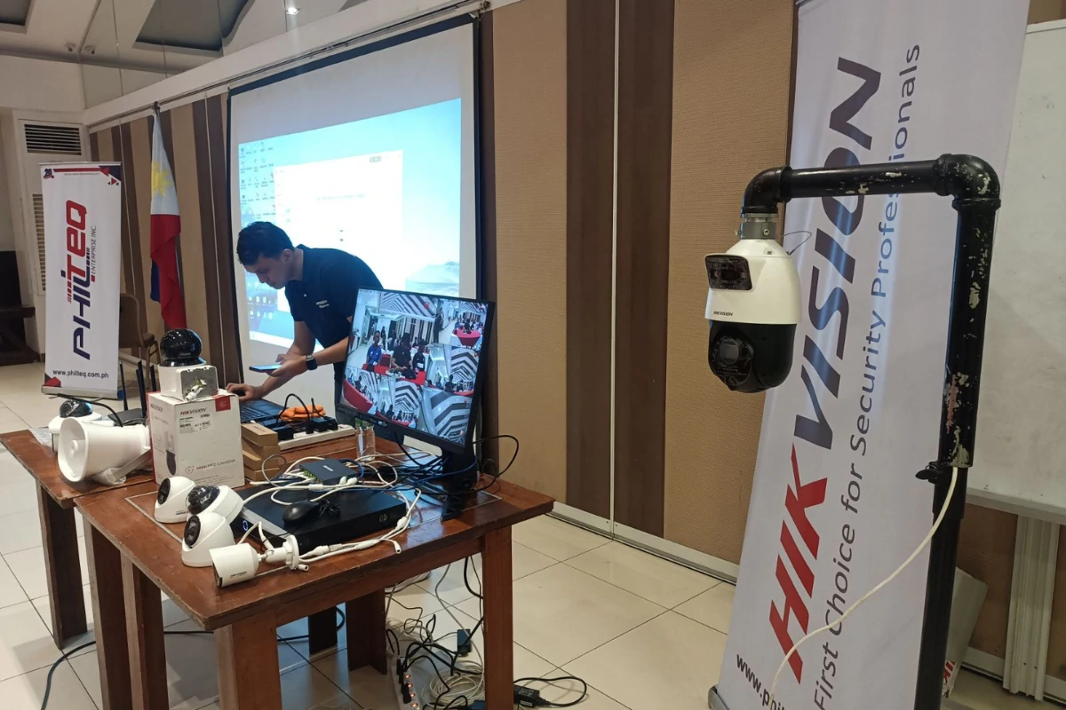 A man adjusts equipment on a table with CCTV cameras and monitors. A screen displays a setup menu. Banners display "PHITED" and "HIKVISION." The setting is professional and technical.