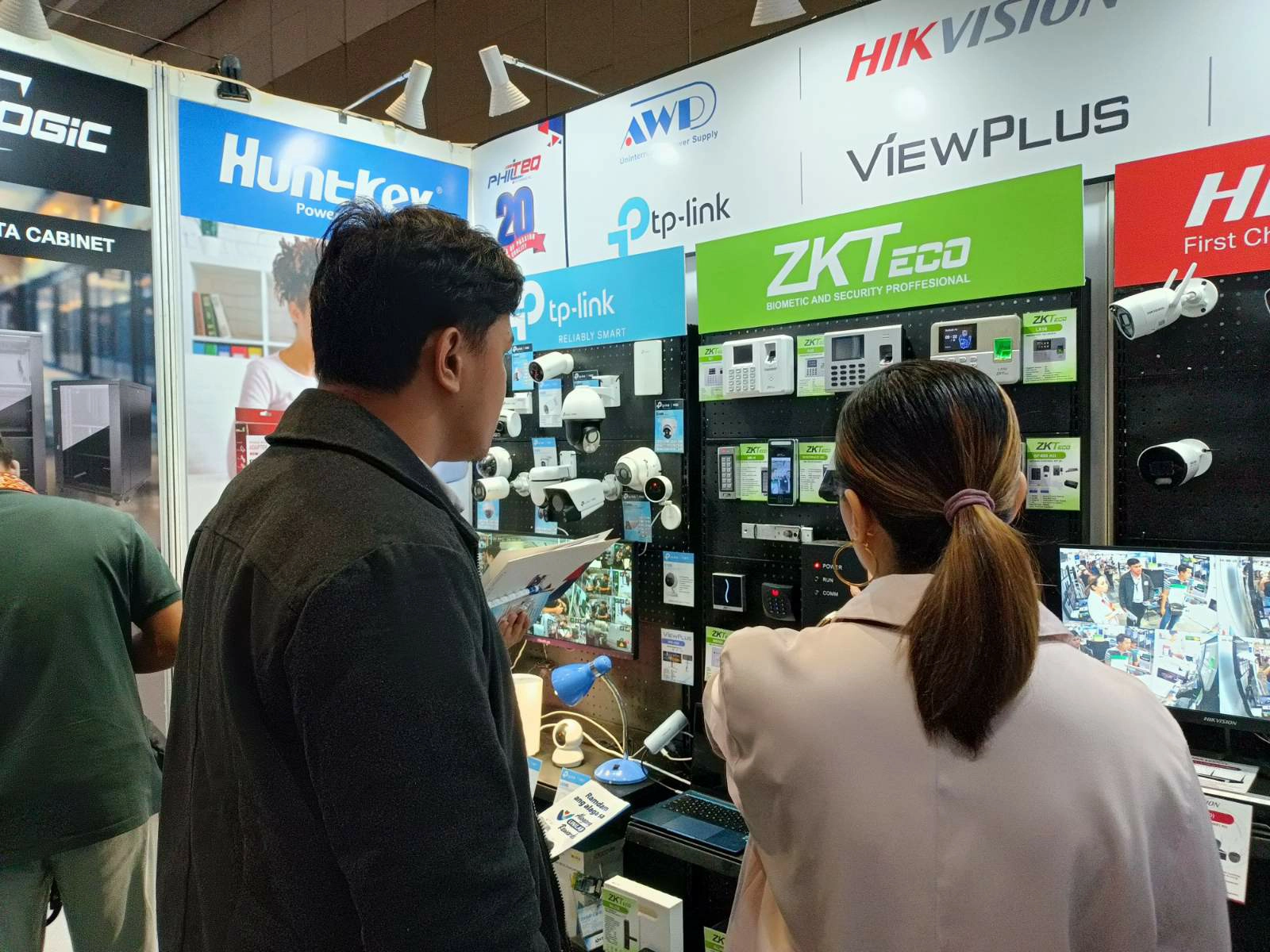 Two people browsing security technology displays in a tech exhibition. Various brands and surveillance cameras are showcased on the walls.