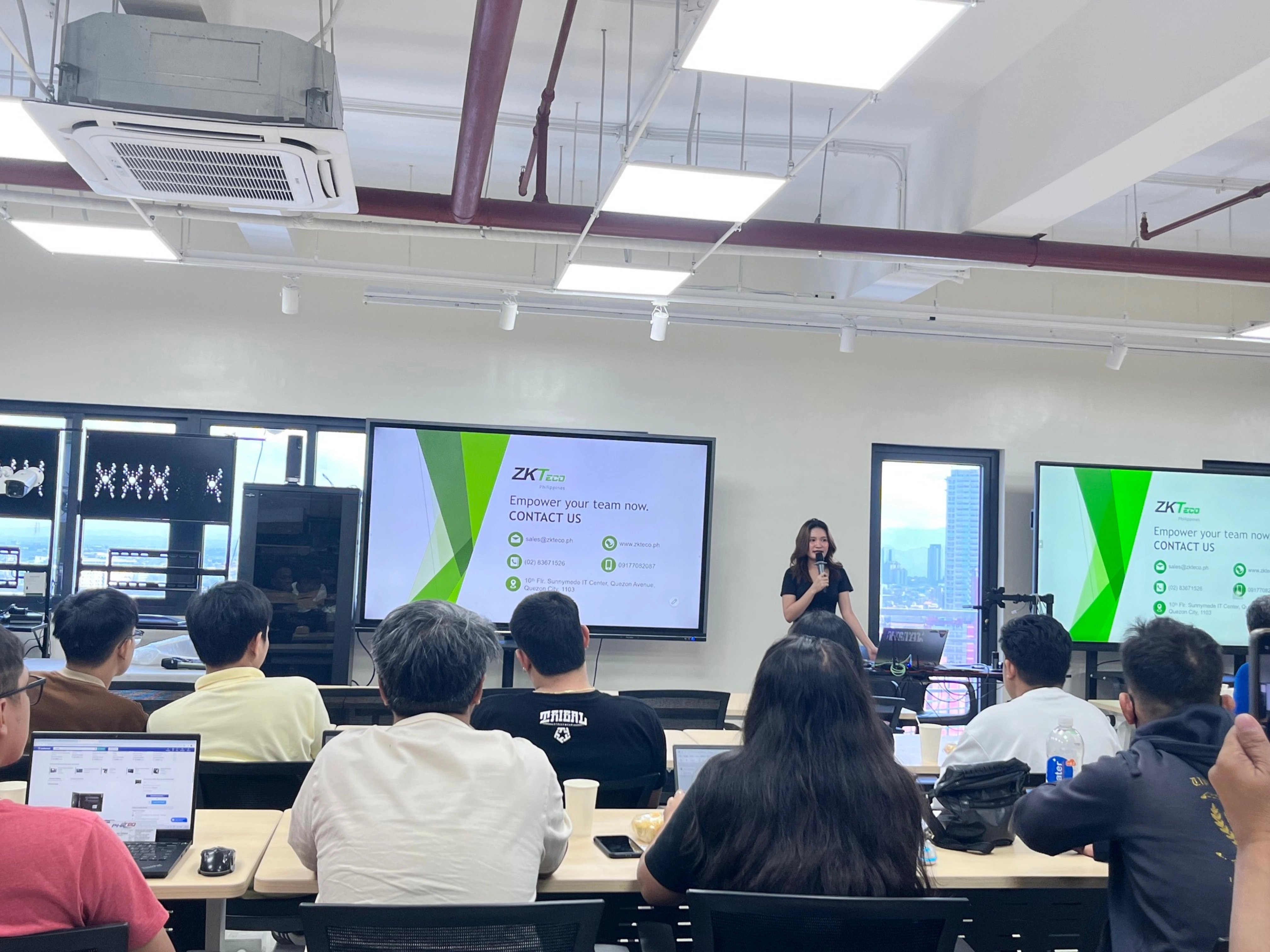 A woman presents to an attentive audience in a modern seminar room. Two large screens display a slide titled "Empower your team now. CONTACT US."