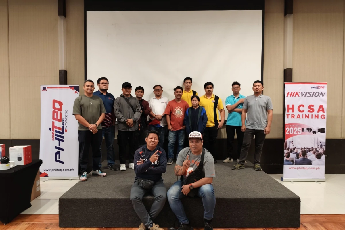 A group of fifteen people, mostly men, stand and kneel on a stage in a conference room. Two banners with "Philteq" and "Hikvision HCSA Training 2023" flank the group. The mood is casual and positive.