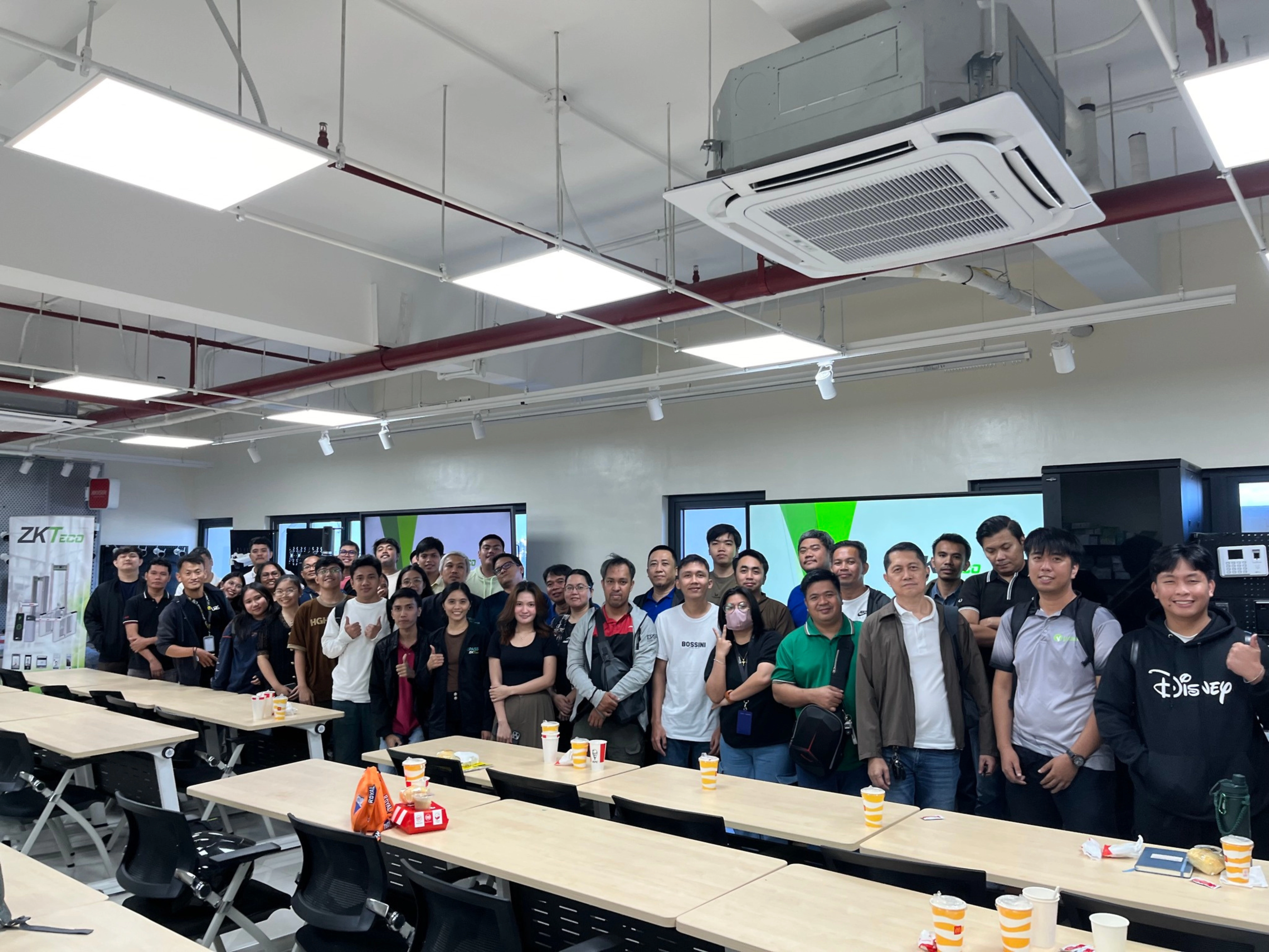 Group of people in a brightly lit classroom stand smiling in front of long tables with chairs. Air conditioning units and screens are visible. Energetic and casual mood.