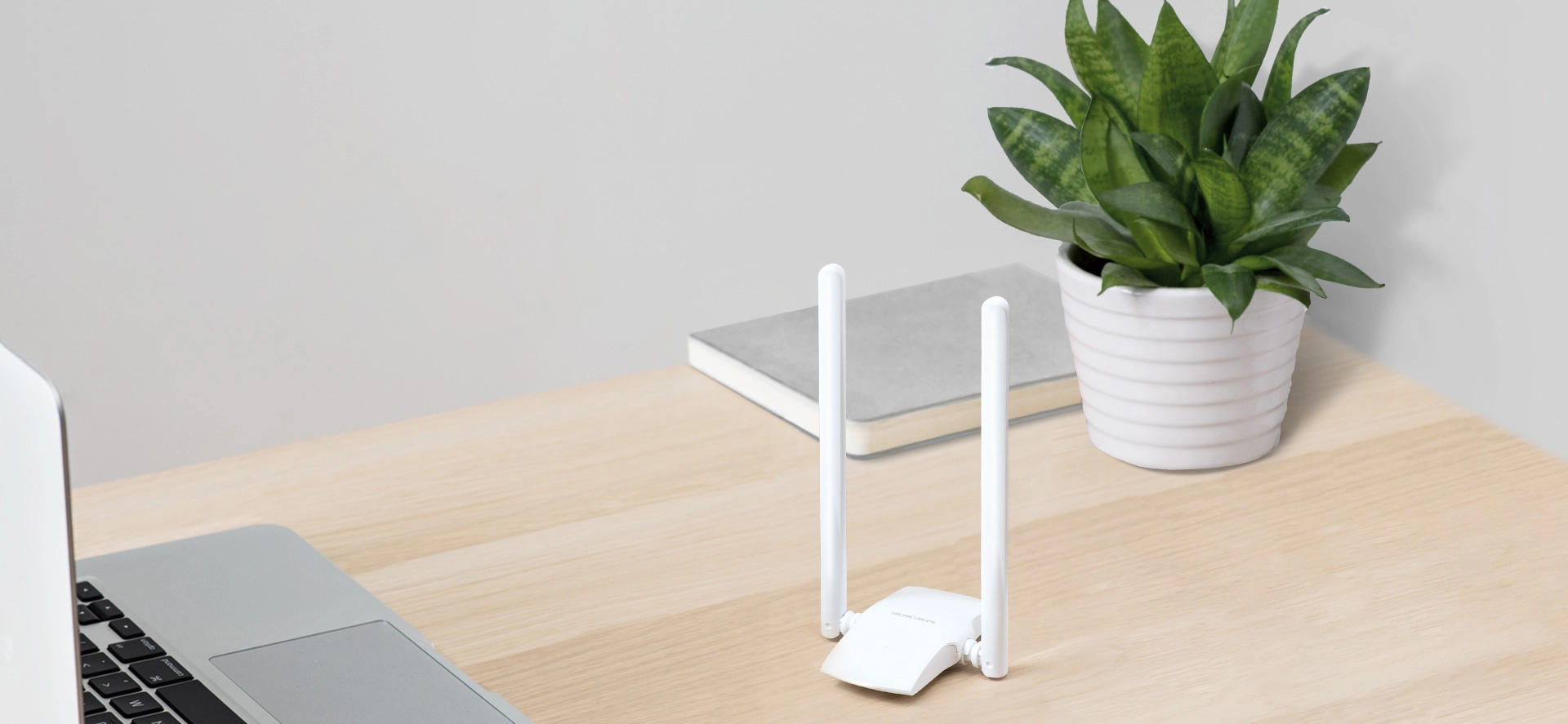 A tidy desk with a laptop, a white Wi-Fi router with two antennas, a potted green plant, and a closed notebook, conveying a minimalist and organized workspace.