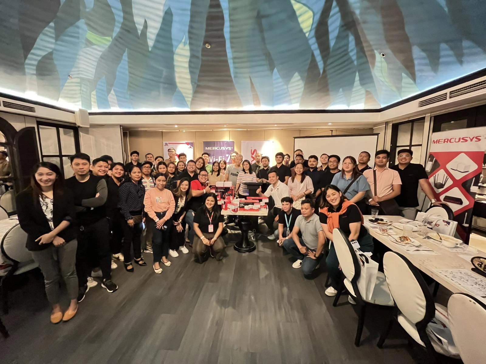 A large group of people smile while posing in a well-lit room with a decorative ceiling. They're gathered around a table displaying tech products.