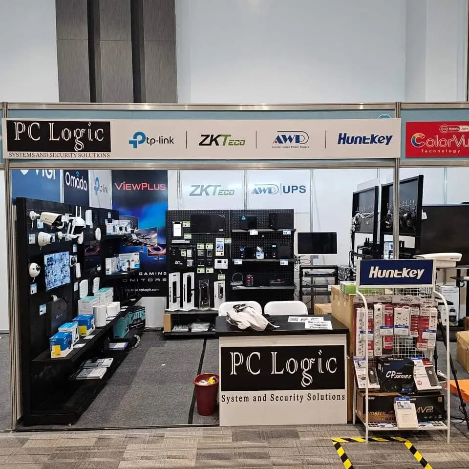 Exhibition booth for PC Logic showcasing security systems and electronics. The setup includes products from brands like tp-link and Huntkey.
